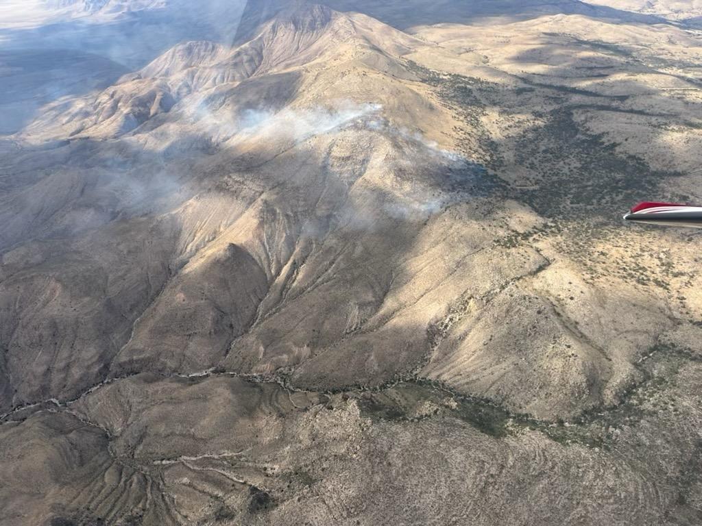 An aerial image looking down on a rocky, sparsely vegetated landscape. Smoke is wafting up from the top and sides of the mountain covered in brown vegetation that is within view. 