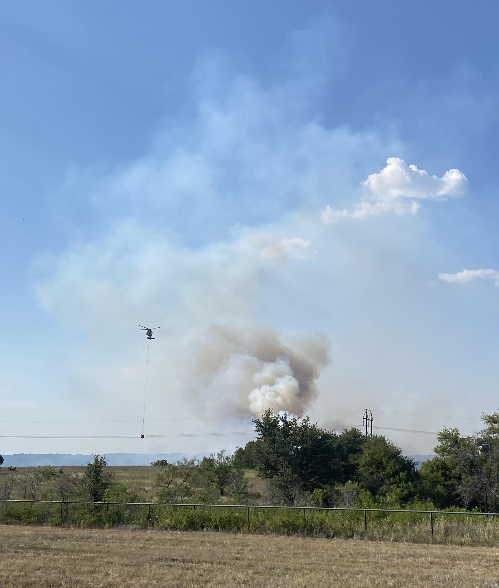 A small black helmet can be seen flying in front of a large gray and white smoke column, rising up from thick green brush burning. 