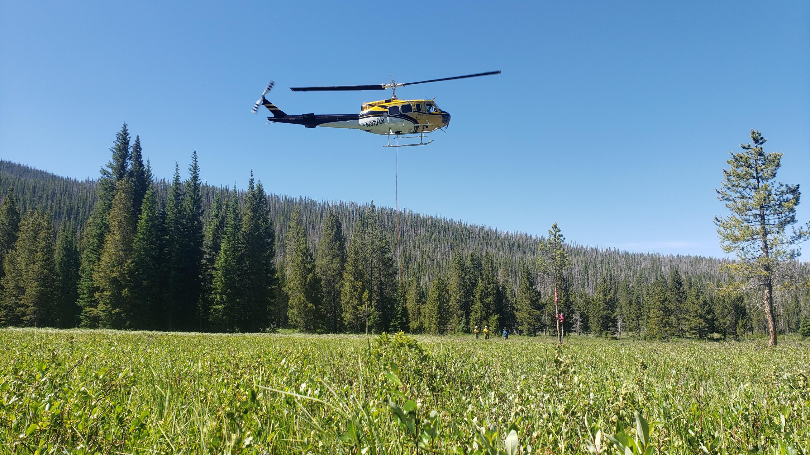 Picture shows a Bell 205 helicopter in the air. Flying over a green meadow with firefighters in the field below. 