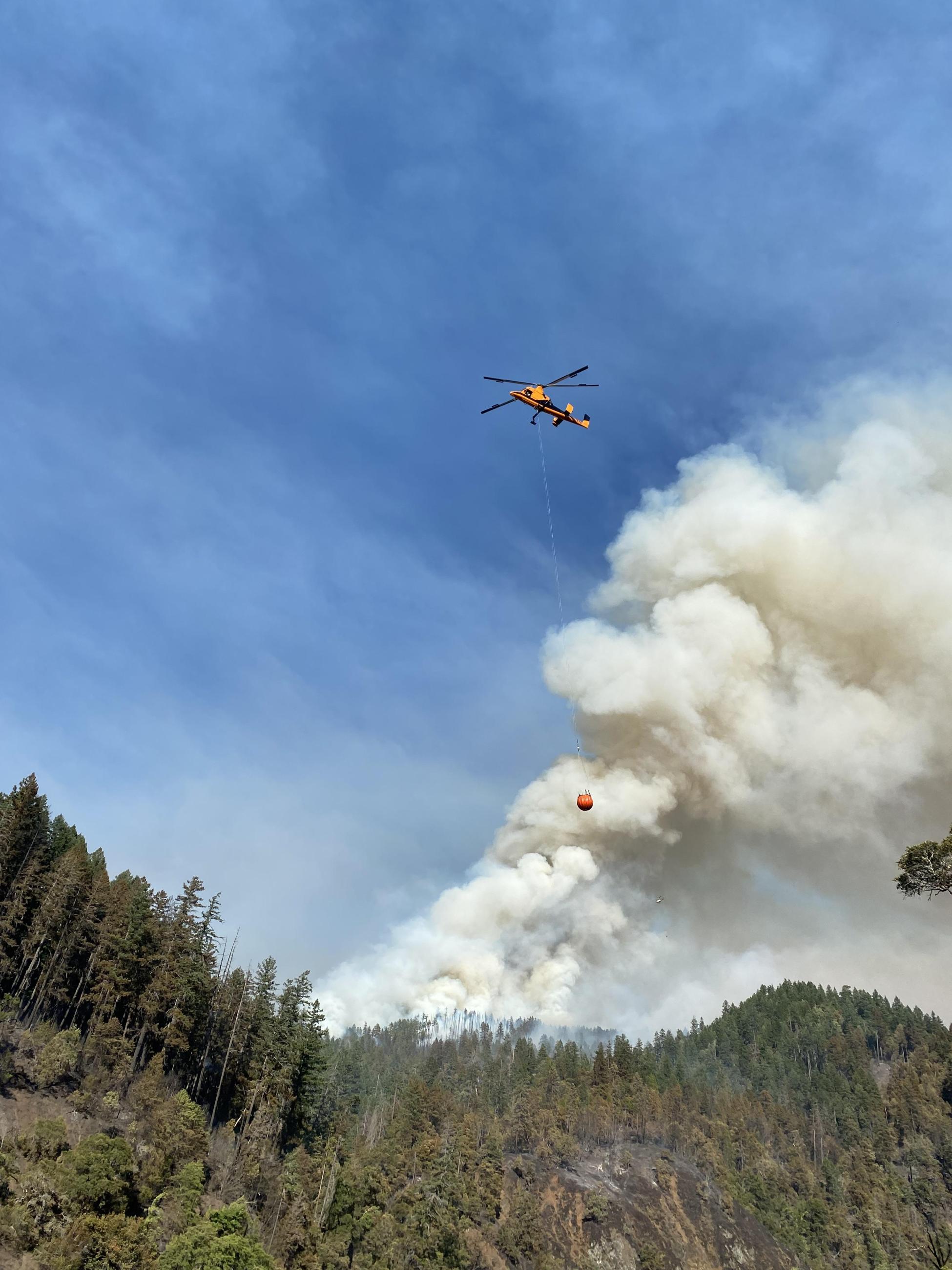 A K-Max helicopter with a bucket attached flies over the Flat Fire. A smoke plume can be seen in the background.