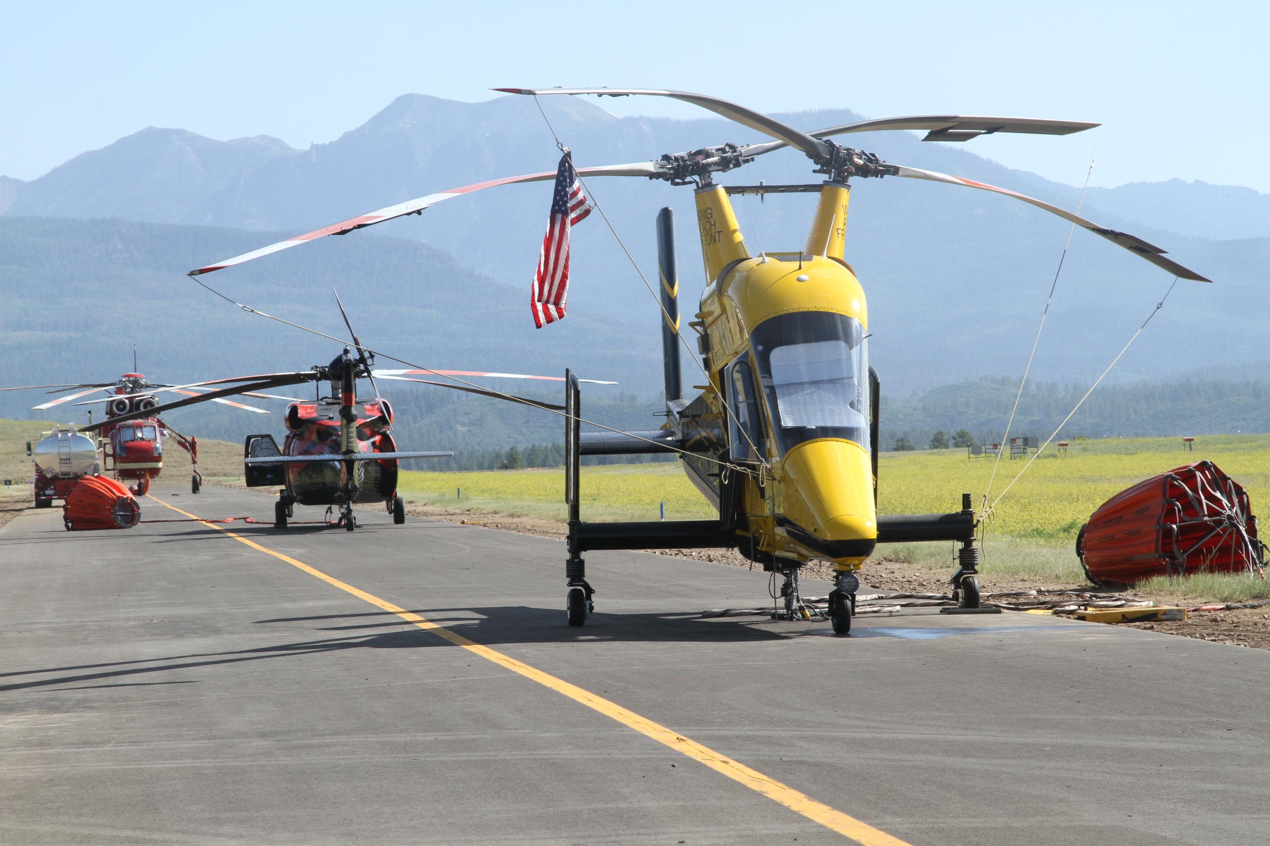 Yellow helicopter sits in front of two red helicopters on a runway.The Rocky Mountains are behind all three helicopters