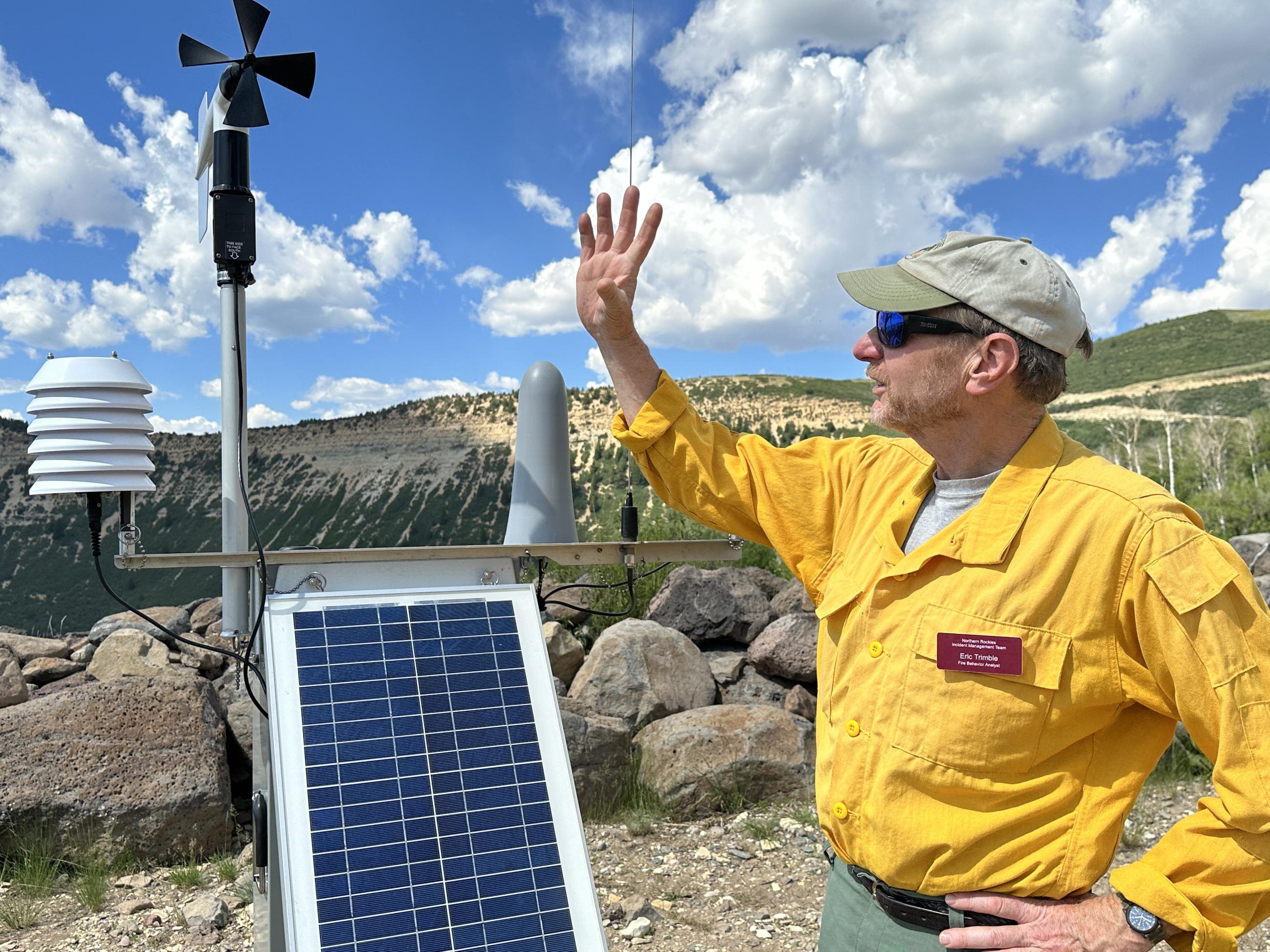 a fire behavior analyst stands next to a weather station on a mountain near the fire