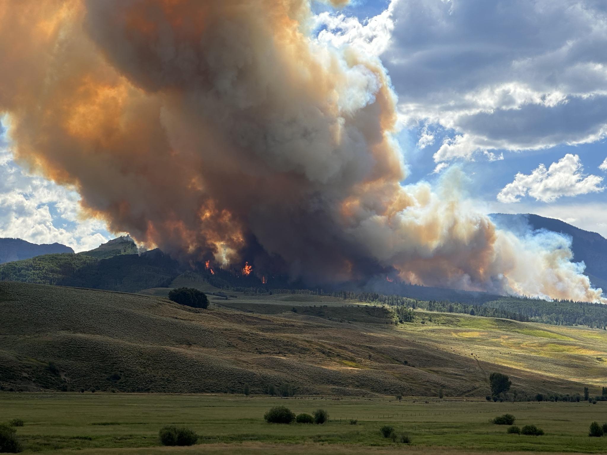 Large flames and heavy smoke in the near distance along a hillside. Green and brown hues on the hilly ground with a few small patches sagebrush in the forefront.