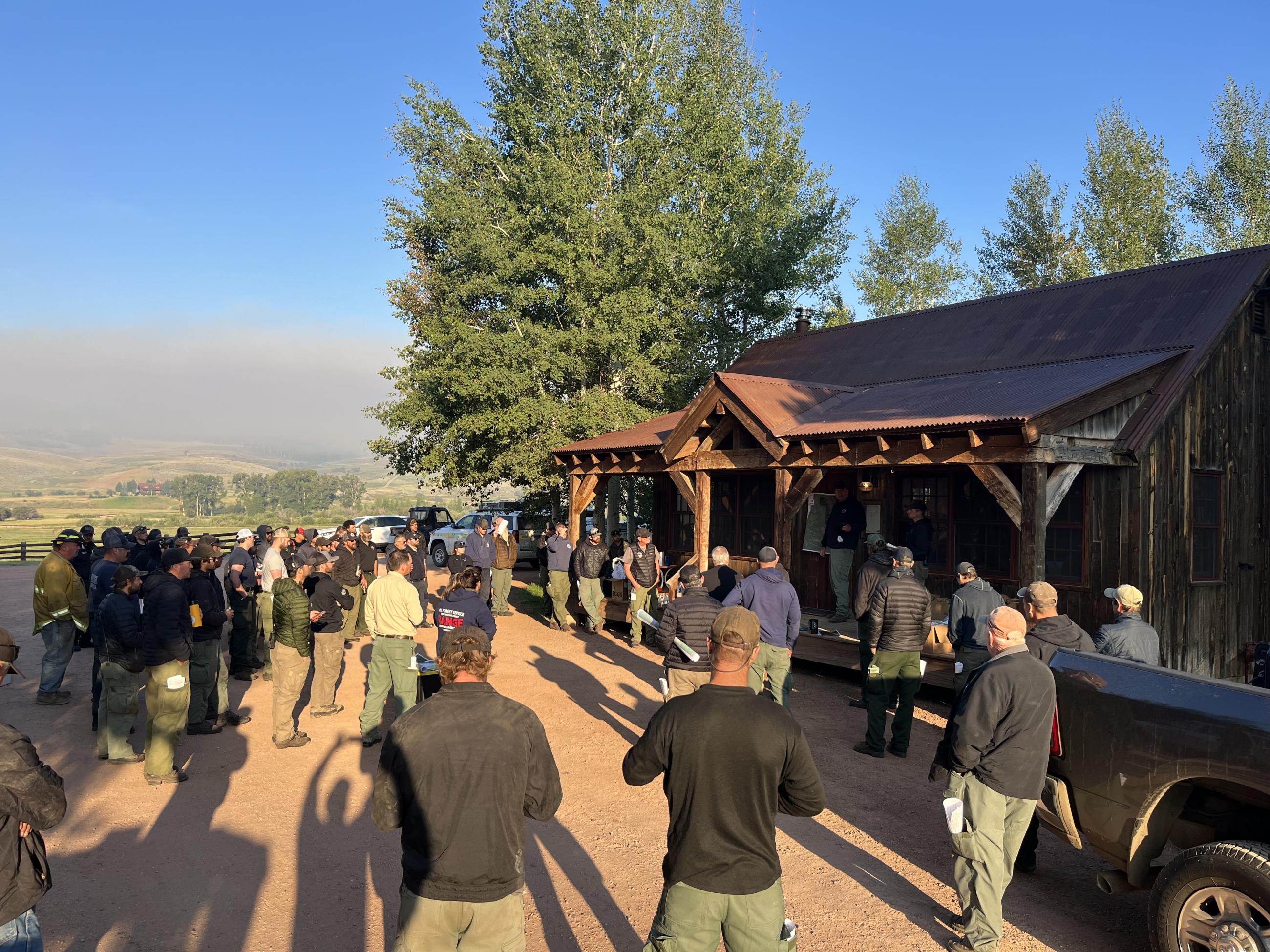 A large group of firefighters standing outside of a cabin as they listen to an operational briefing for the day.