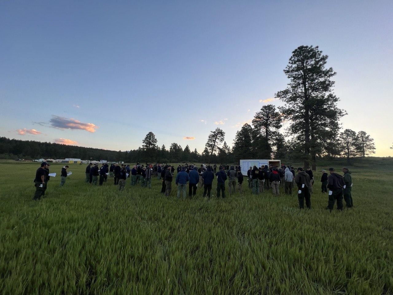 large group of people standing in green grass field as the sun rises in a mostly clear blue sky. People are surrounding a trailer with a map tape on the side to get a morning briefing