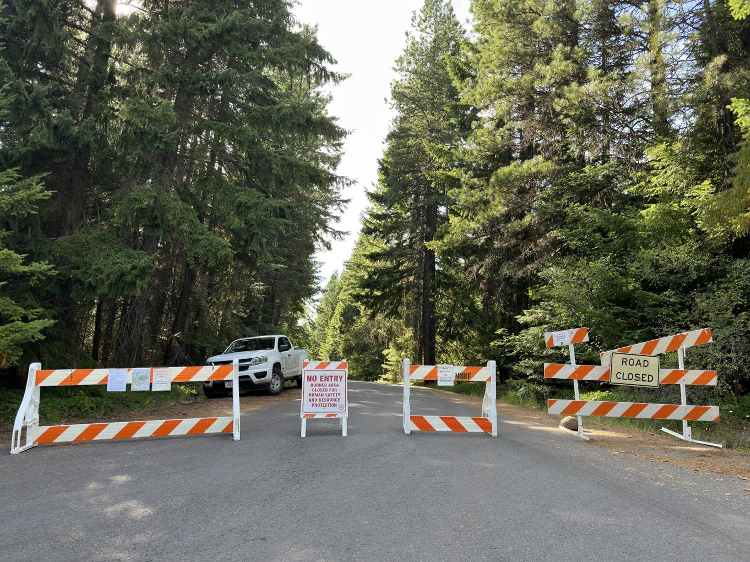 Image shows road closure signs and road barriers across the end of a paved and tree-lined forest road. Also pictured behind the barriers is a road guard's white pick-up truck. 