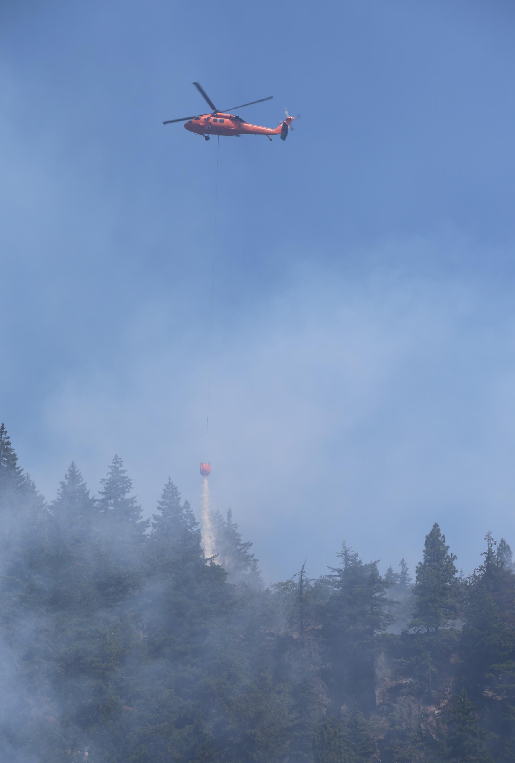 Helicopter dropping water from long-line bucket configuration into smoking forested hillside.