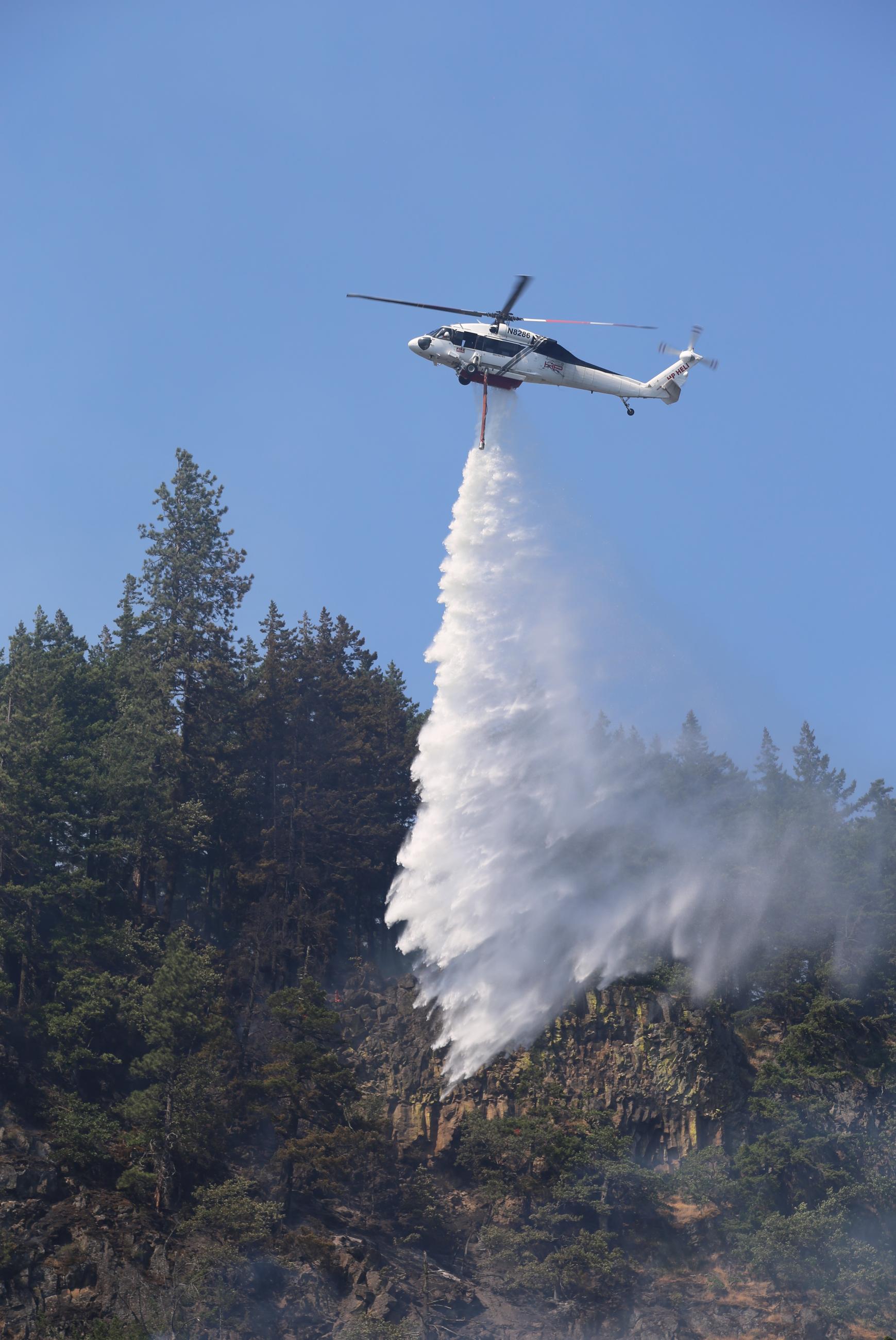 Helicopter dropping water from tank into smoking forested hillside.