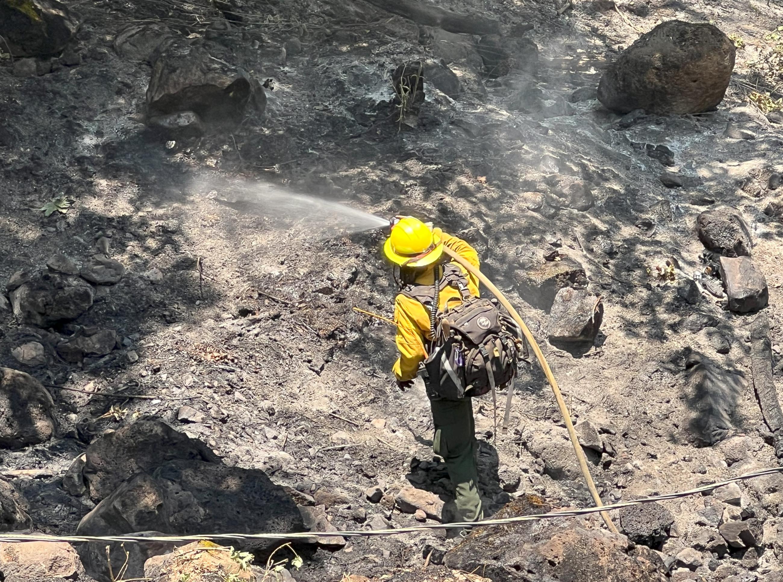 Firefighter sprays water from hose onto smoldering hillside during mop-up operations.