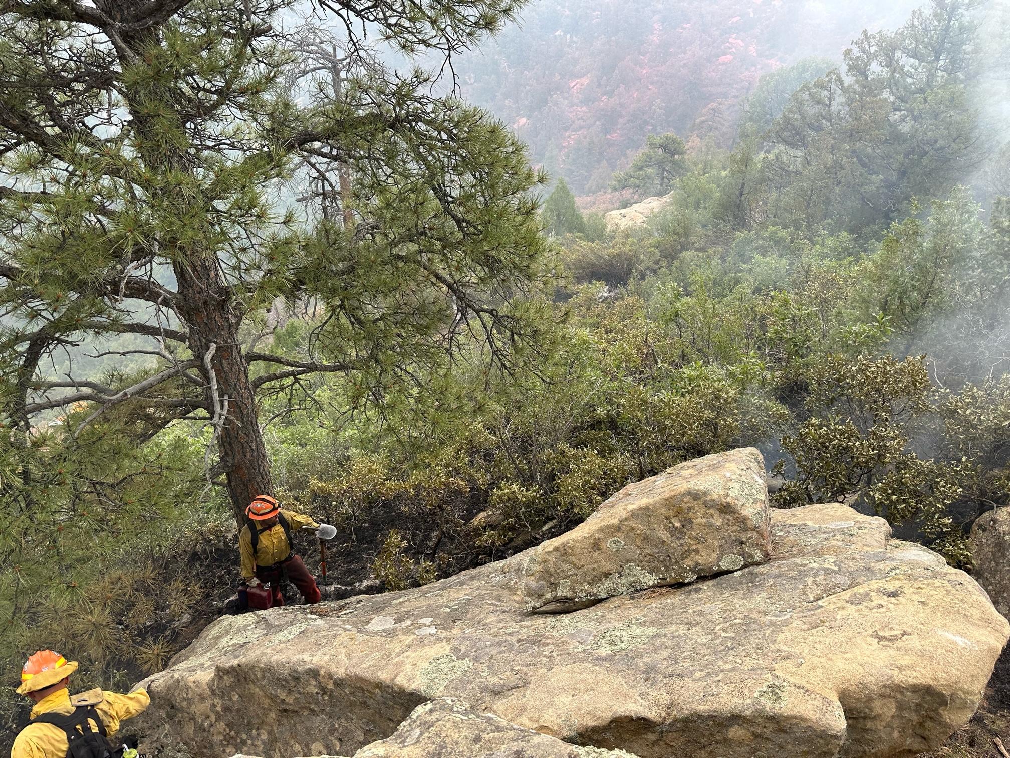Two fire fighters are hiking around a large boulder along the fire line.