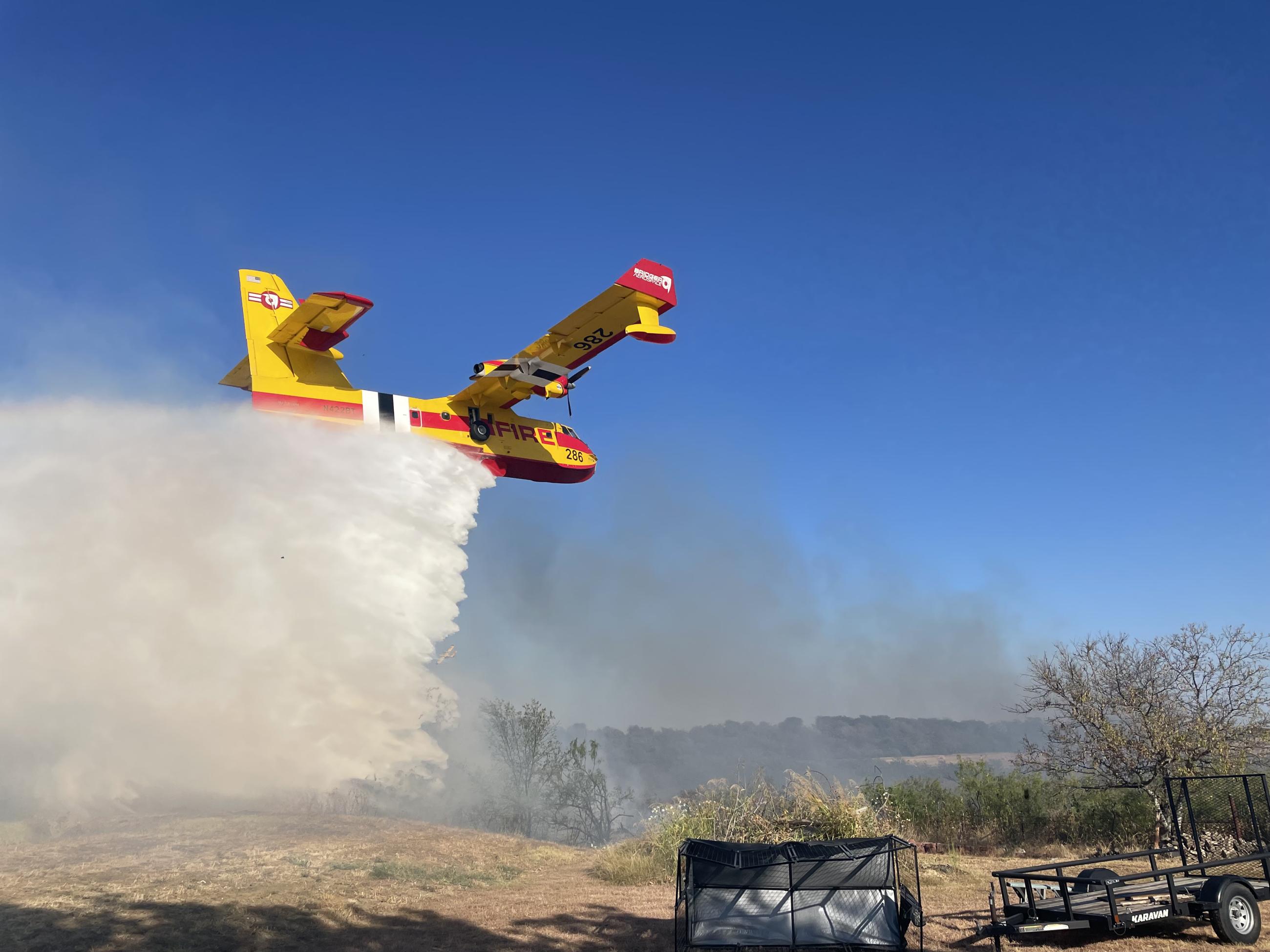 yellow plane flying low to the ground with water dropping from it's belly over black ground from burned grass
