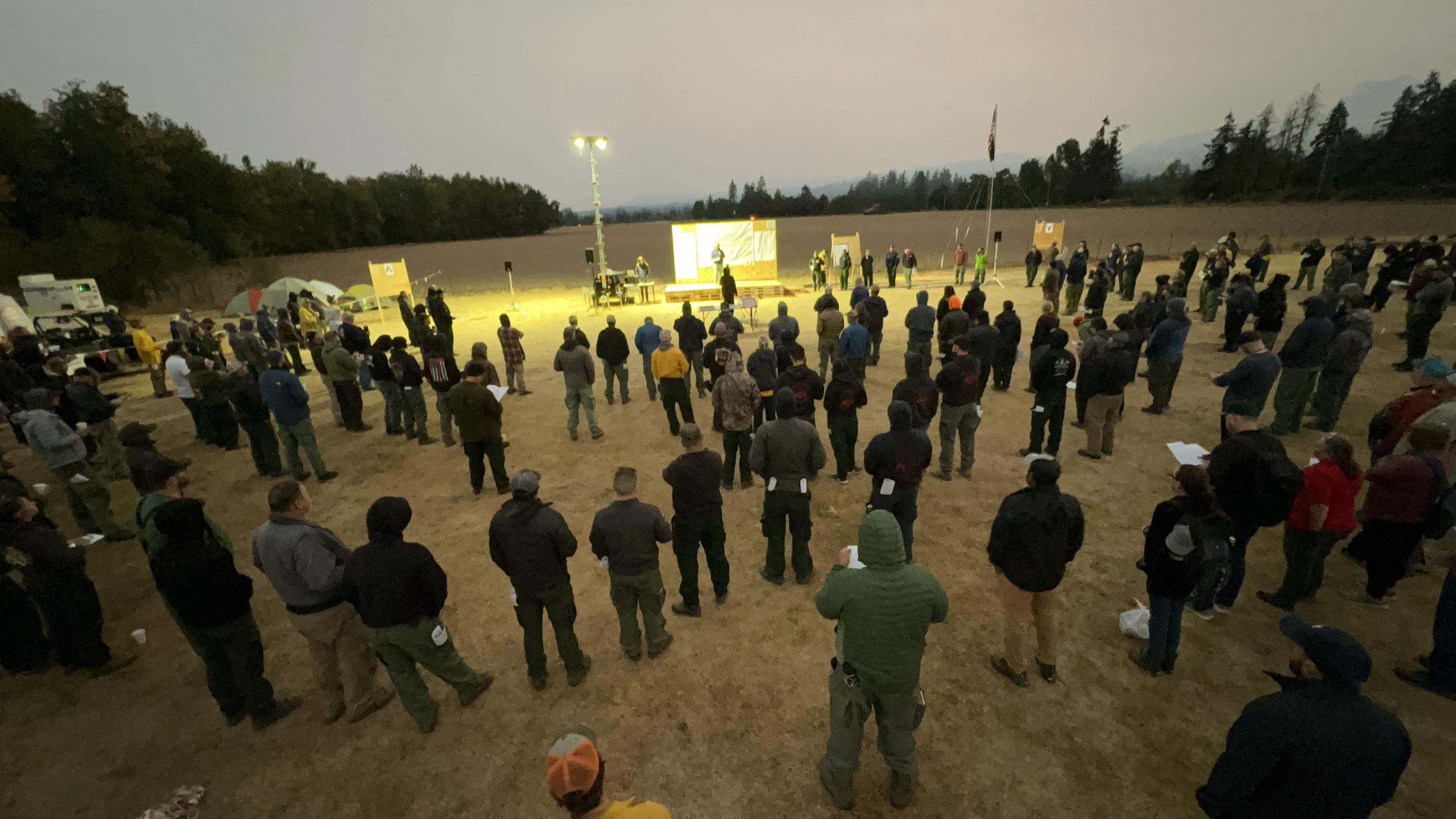 Firefighters are formed in a semi-circle around a bulletin board for a meeting early on August 24, 2023.