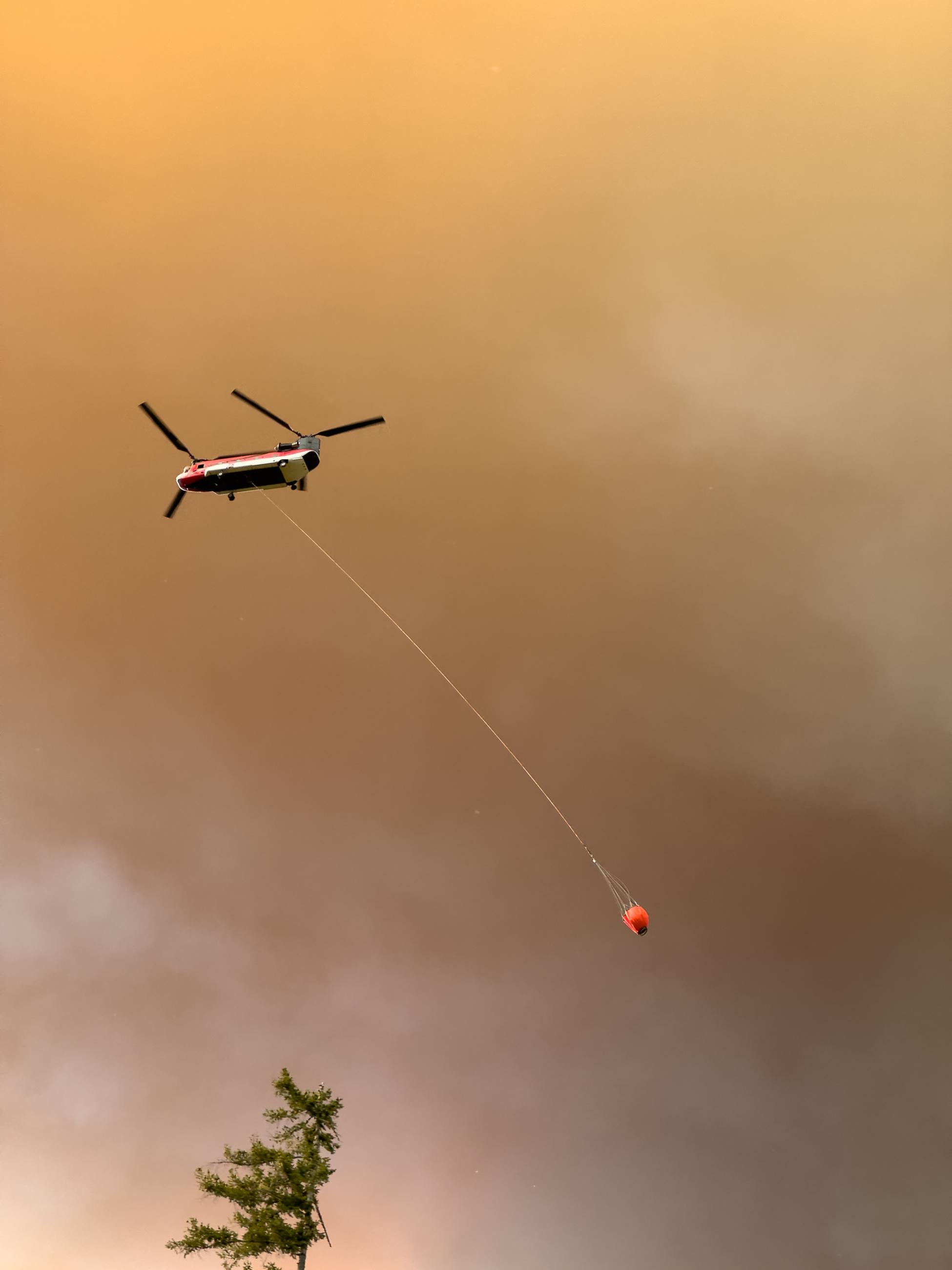 A Chinook helicopter flies below smoke with a water bucket.