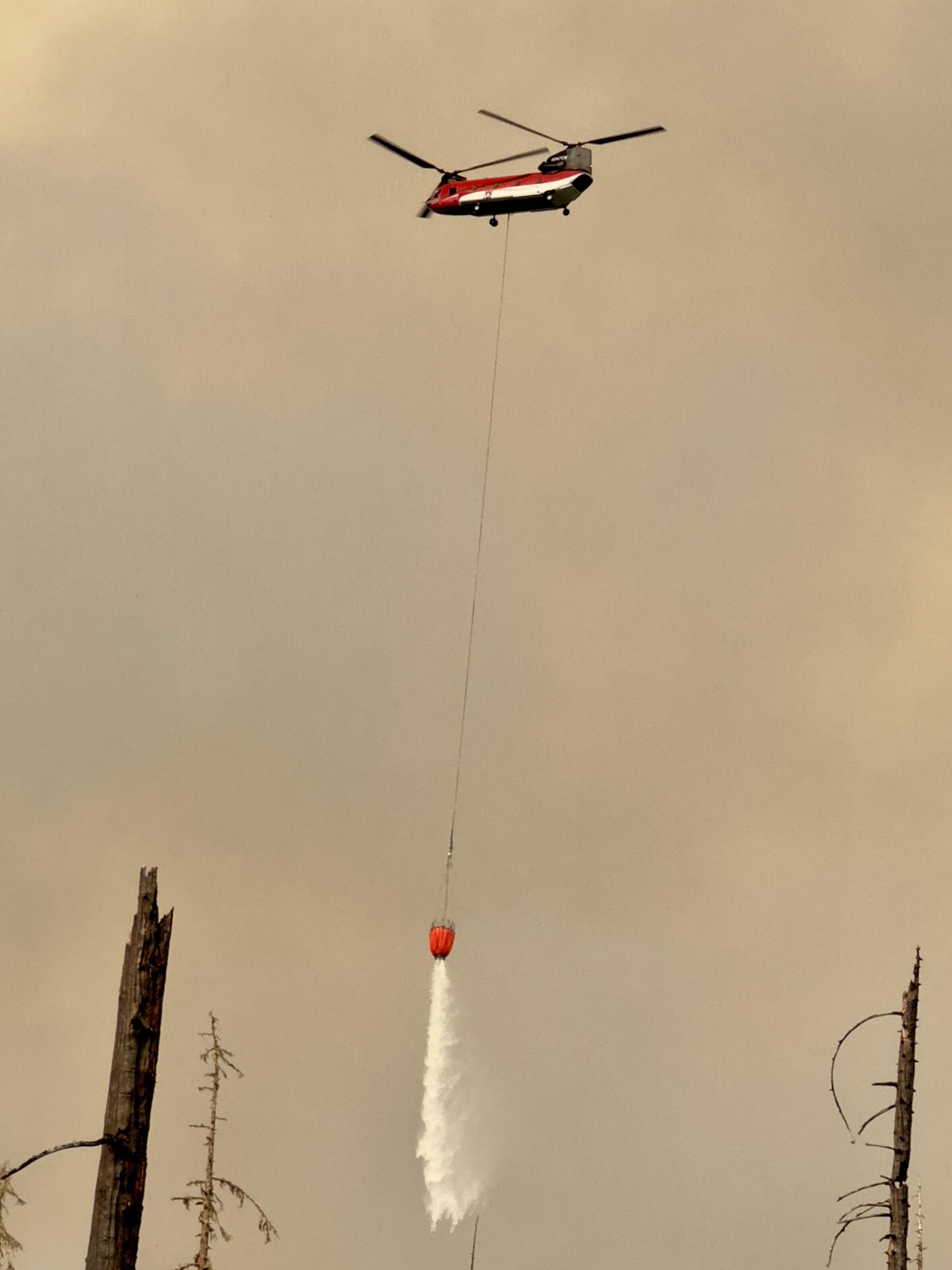 A Chinook helicopter flies below smoky skies with a water bucket that's dumping water.