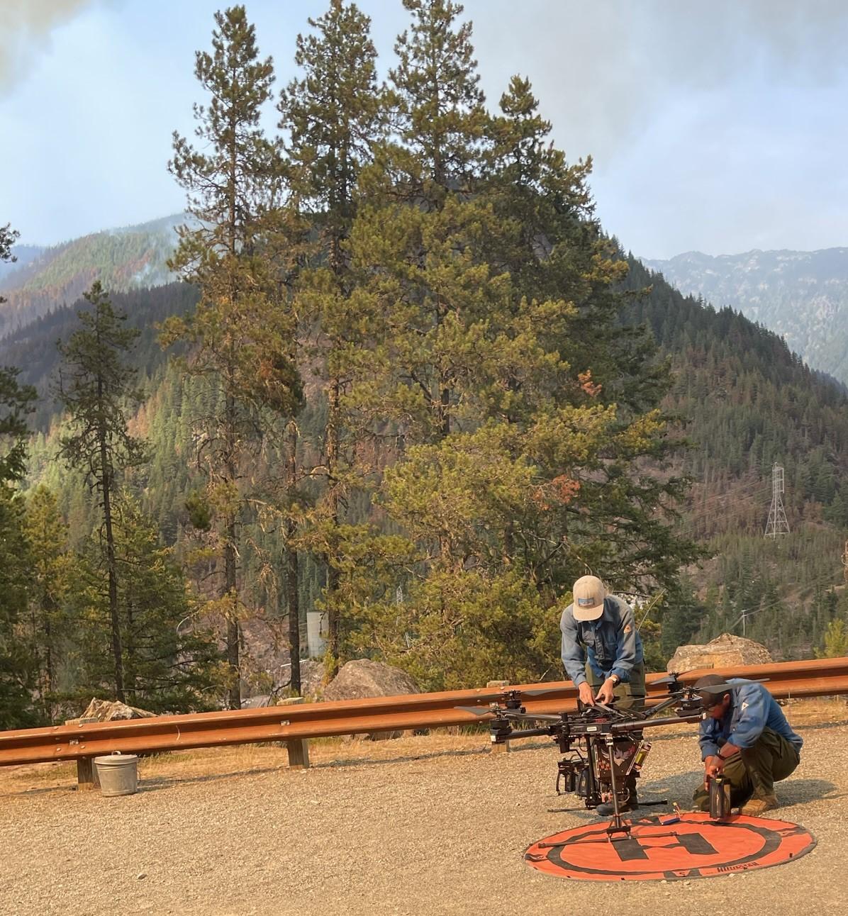 A man and a woman with a unmanned aerial system (UAS or drone) on the side of a mountain highway with trees, a lake, mountains and smoke in the background.