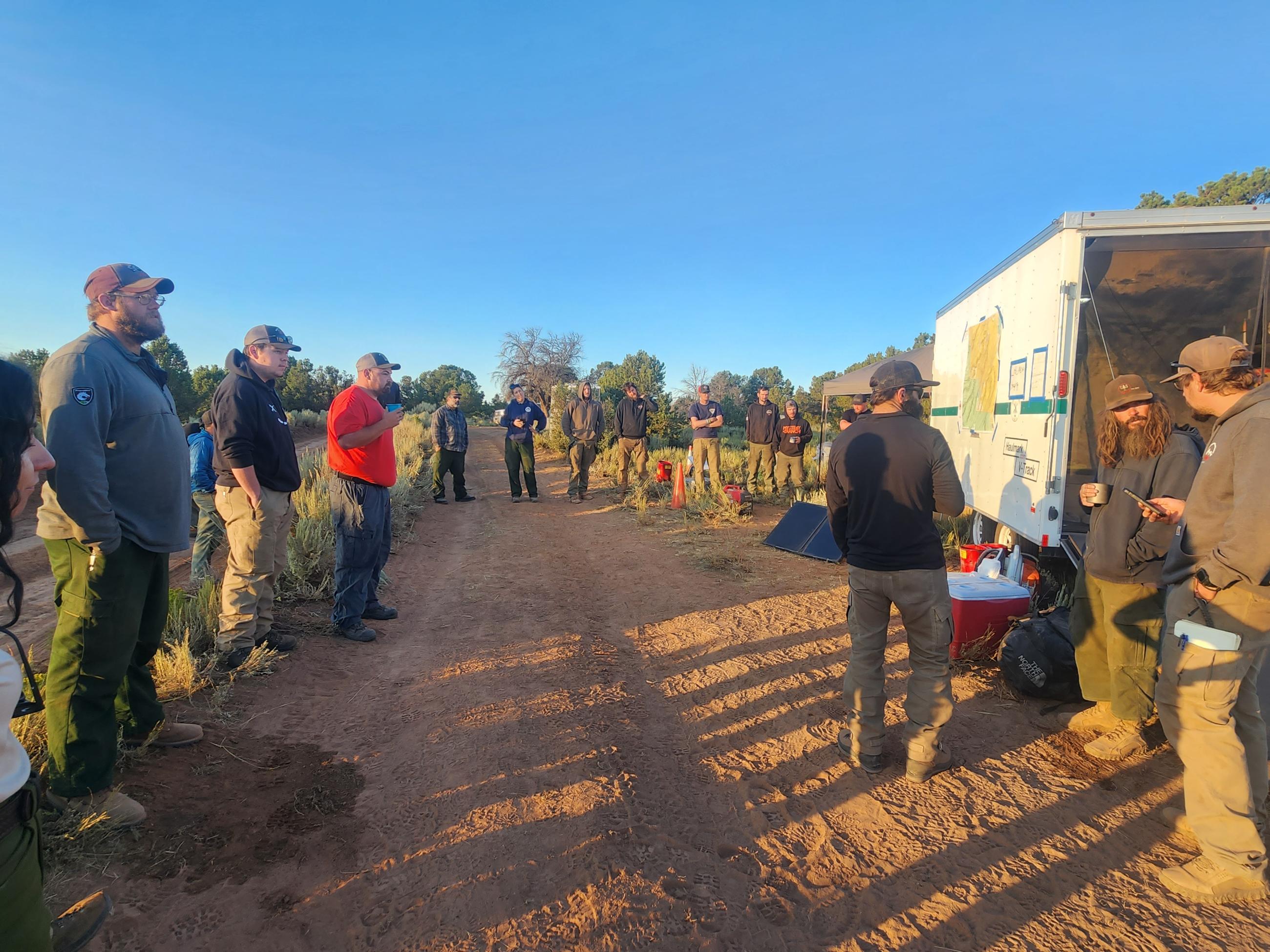 Image of firefighters getting ready for their morning operations briefing on the Little Mesa Fire off a red dirt road with sagebrush. The briefing map it placed on a vehicle for firefighters to view as they hear previous days accomplishments and their assignments for the day.