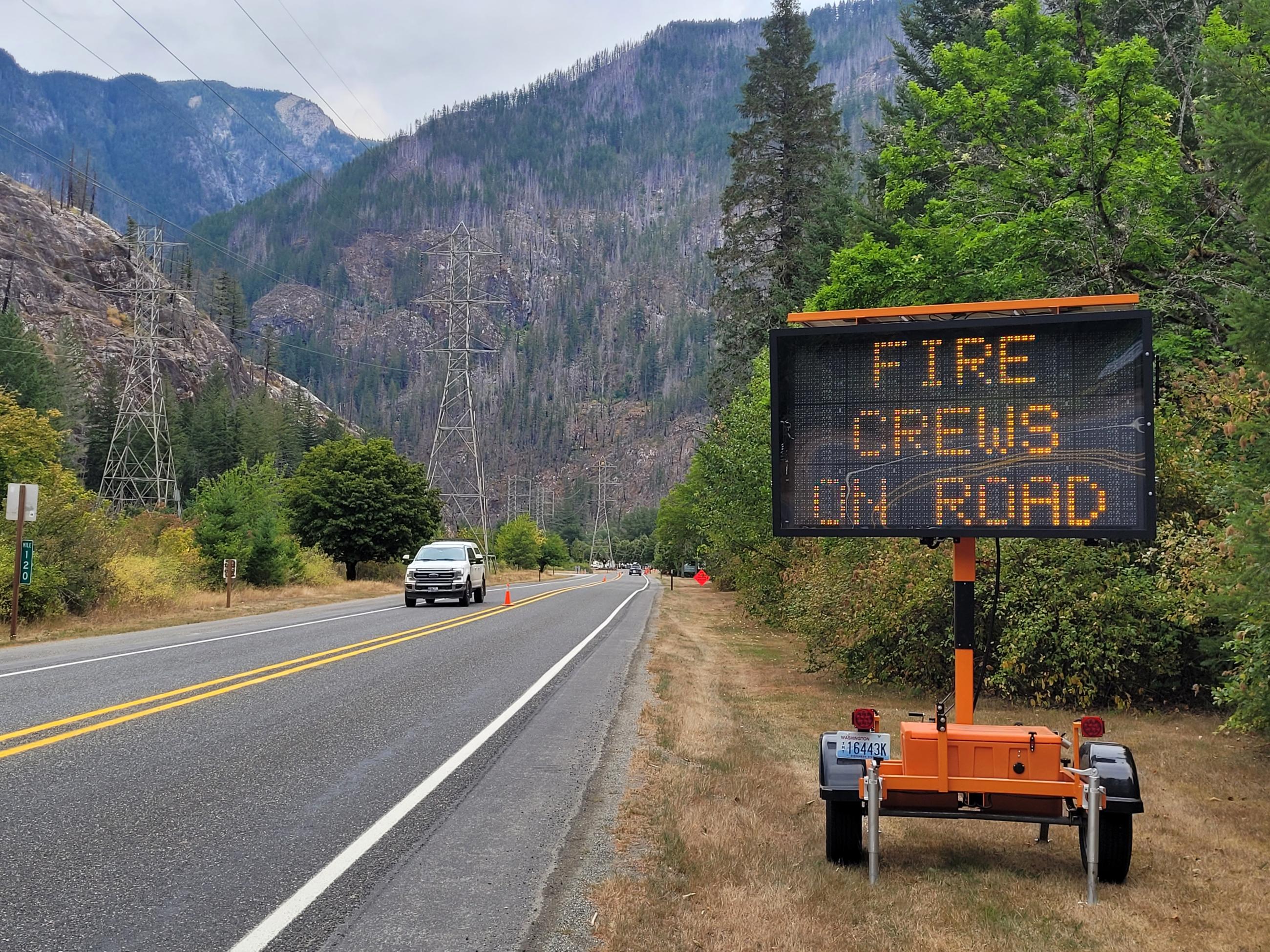 A photo of a large road-side sign with the words "Fire Crews on Road" flashing across while a car drives down the road past the sign. 