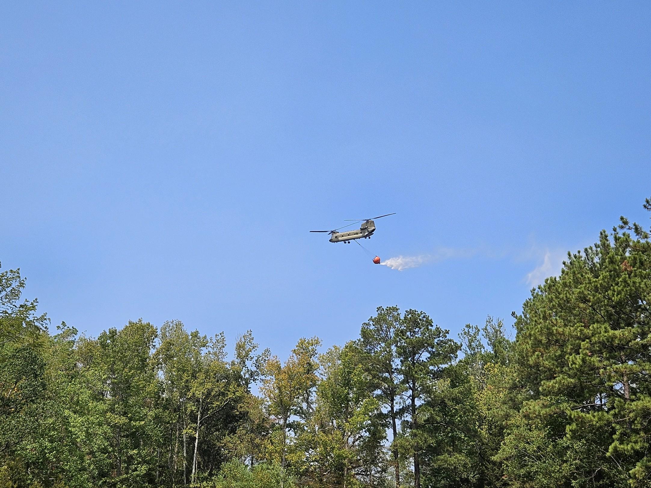 Chinook helicopter carrying a water bucket flies over tree canopy.