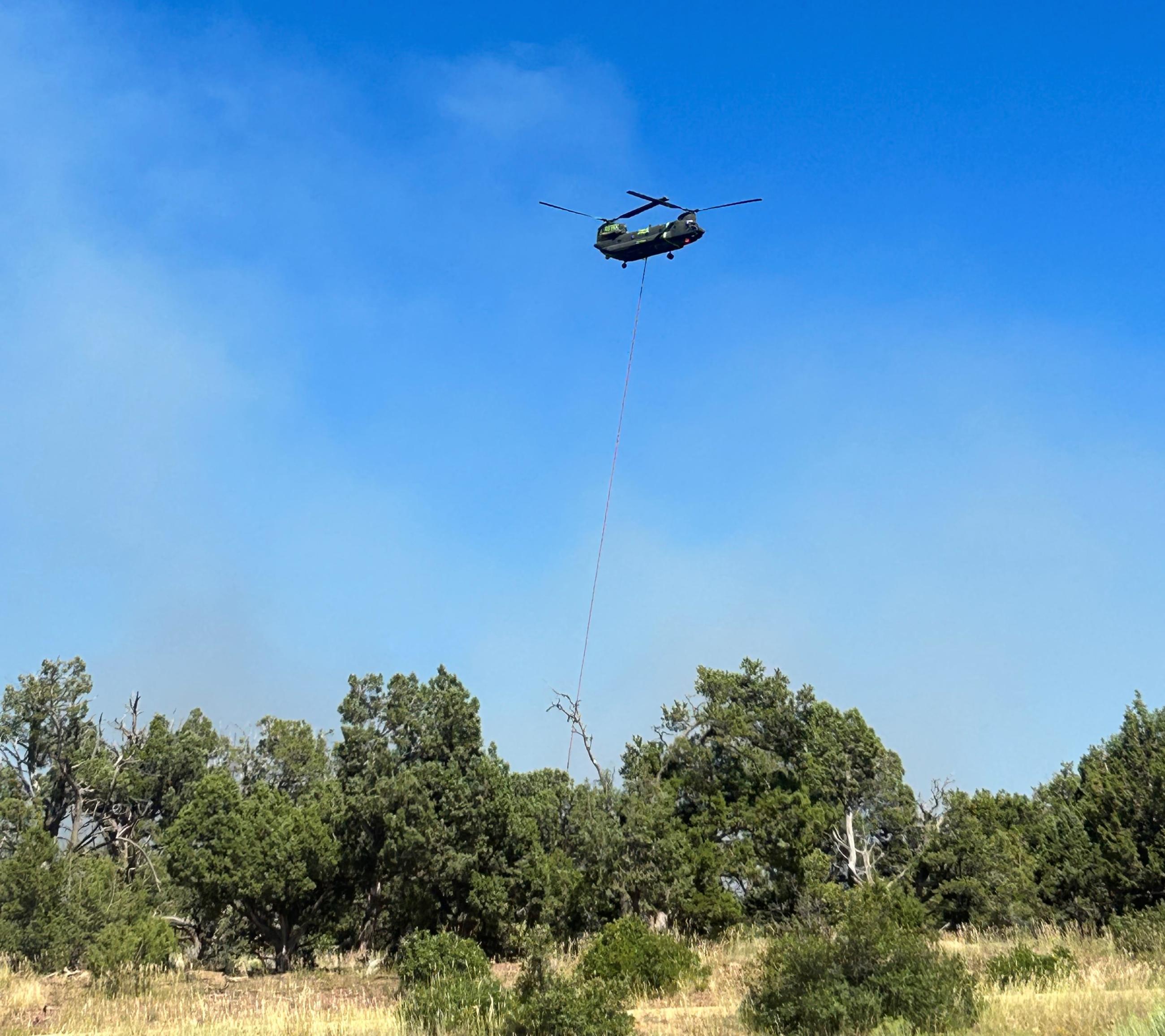 A helicopter drops water over a forested area with light smoke rising