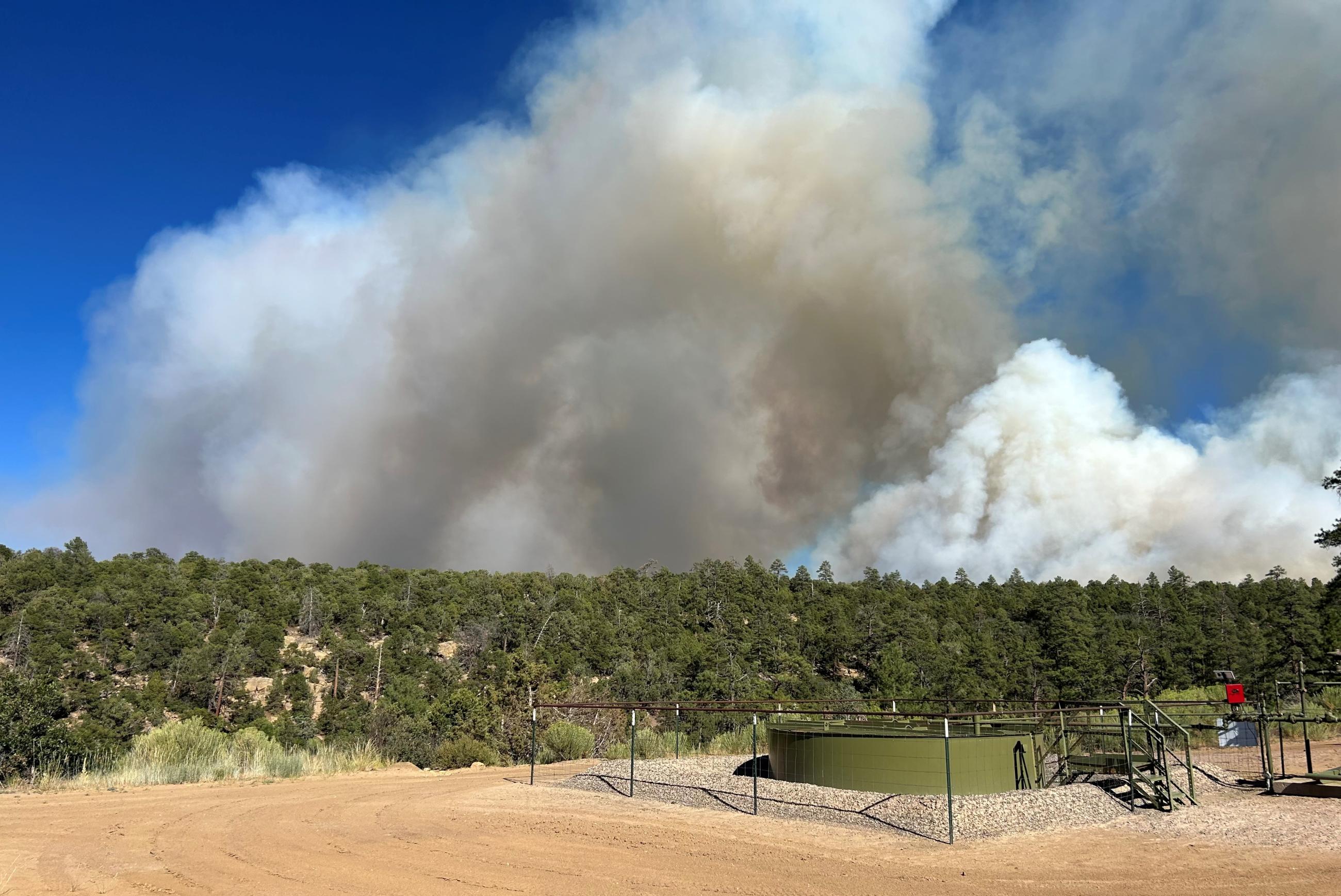 Smoke rises behind a forested hill and natural gas infrastructure.