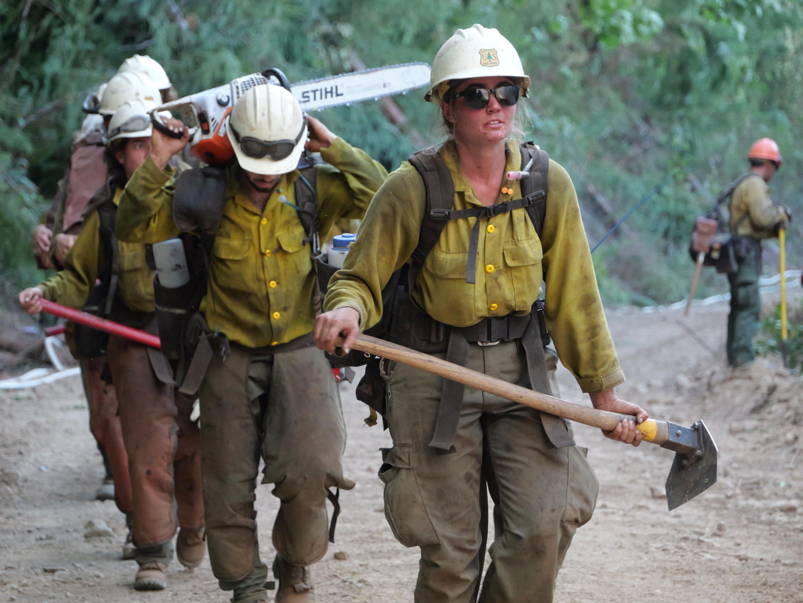 A female firefighter walks at the front of a line of firefighters, all carrying tools, on the Bedrock Fire, Oregon.