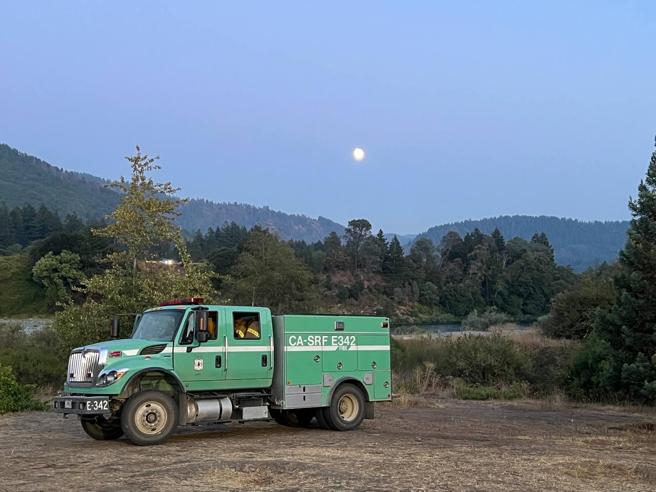 A green forest service fire engine seen at dusk parked on dry grass with a full moon and forested hills in the background