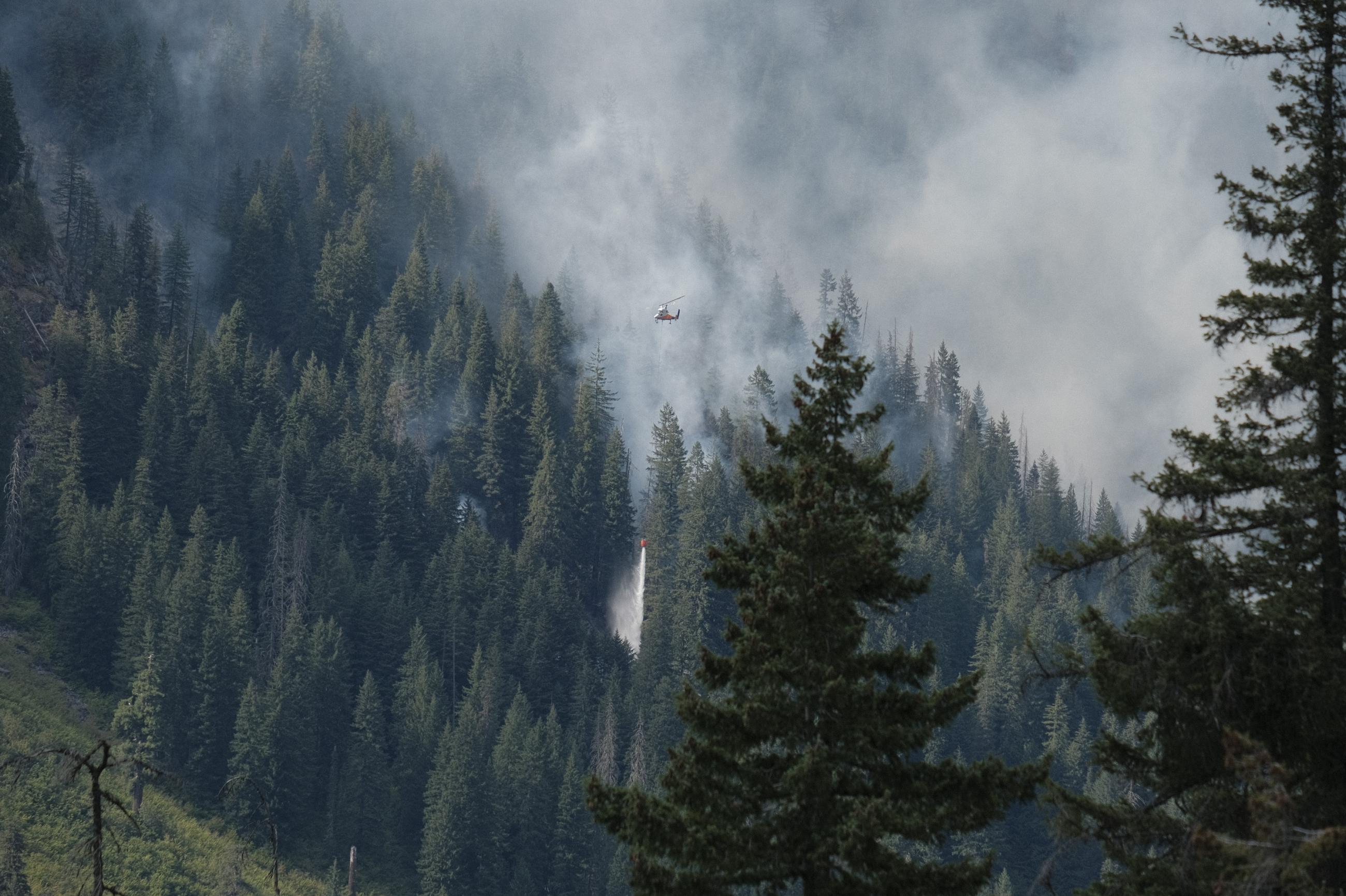 Water is dropped from an orange bucket dangling below a large helicopter on the south flank of the Airplane Lake Fire on August 22, 2023.