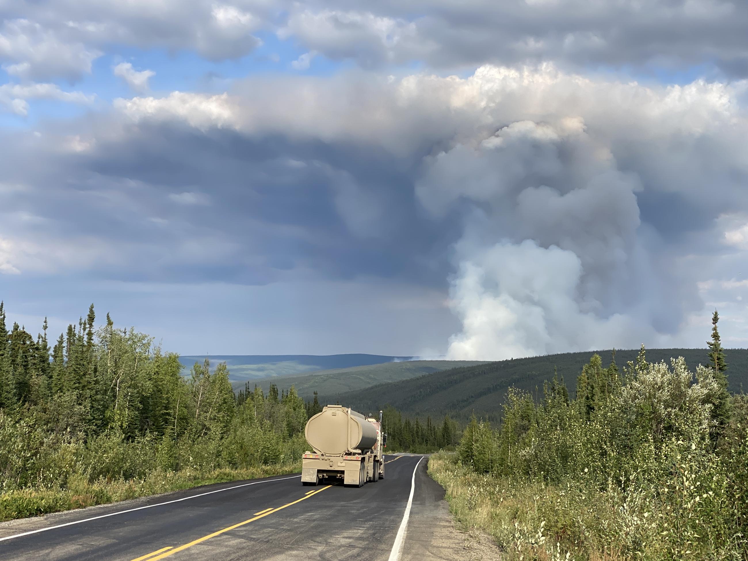 A fire truck drives down a highway with a plume of wildfire smoke.