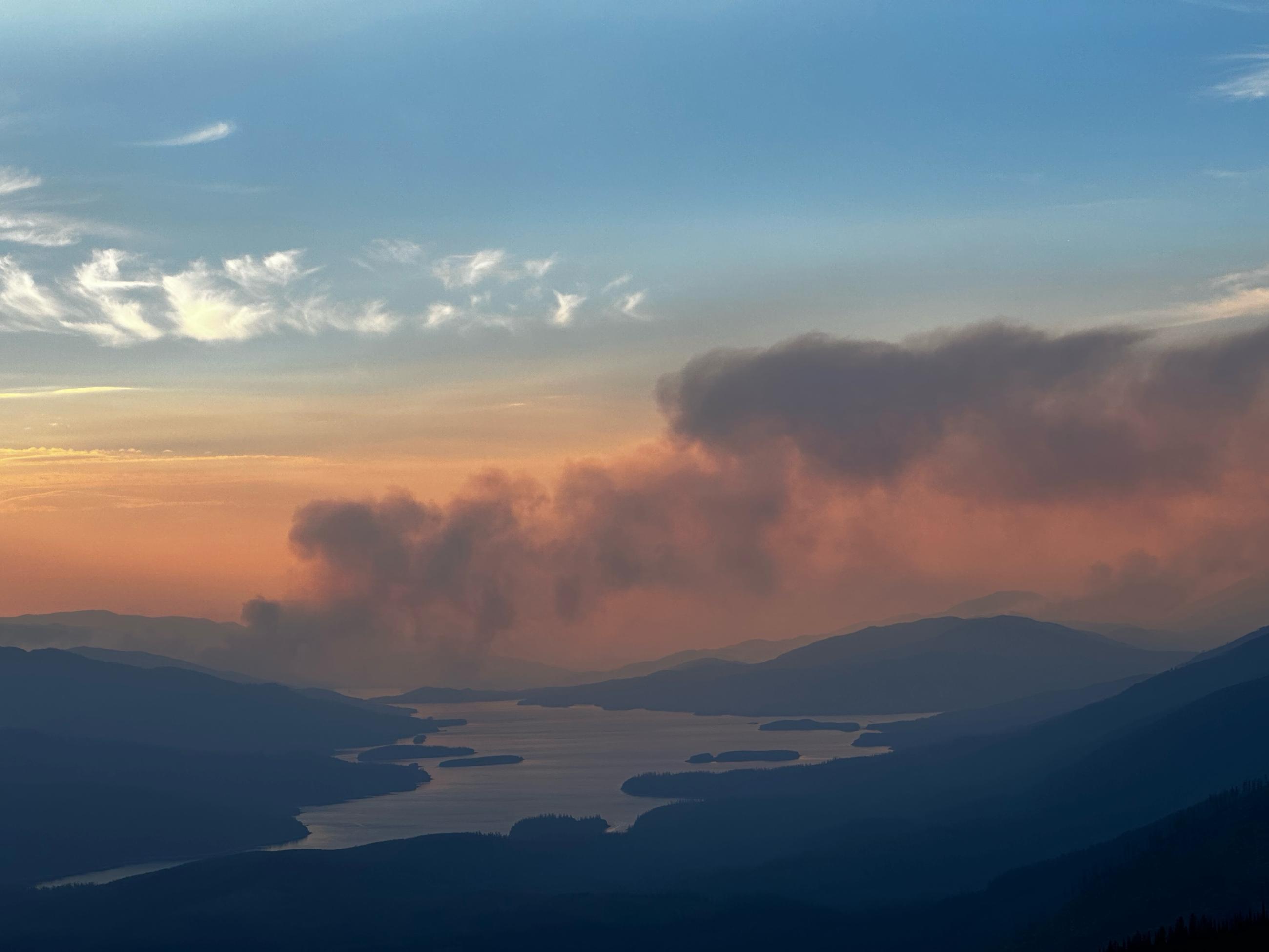 Mountain landscape at sunset with Doris Fire smoke column drifting to the right and Hungry Horse Reservoir down in the middle