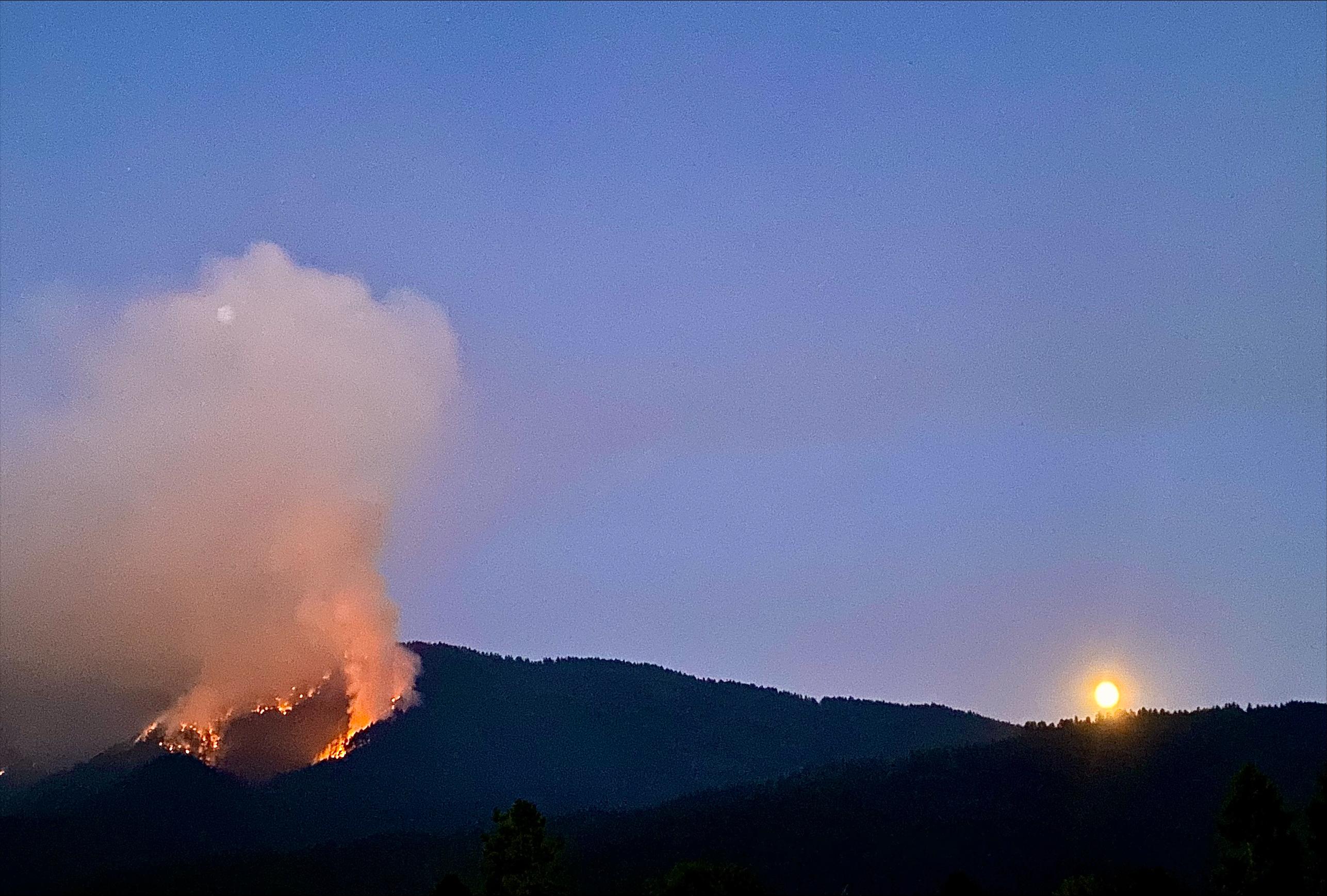 A circle of fire is burning in the mountains at night with a full moon in the backrgound