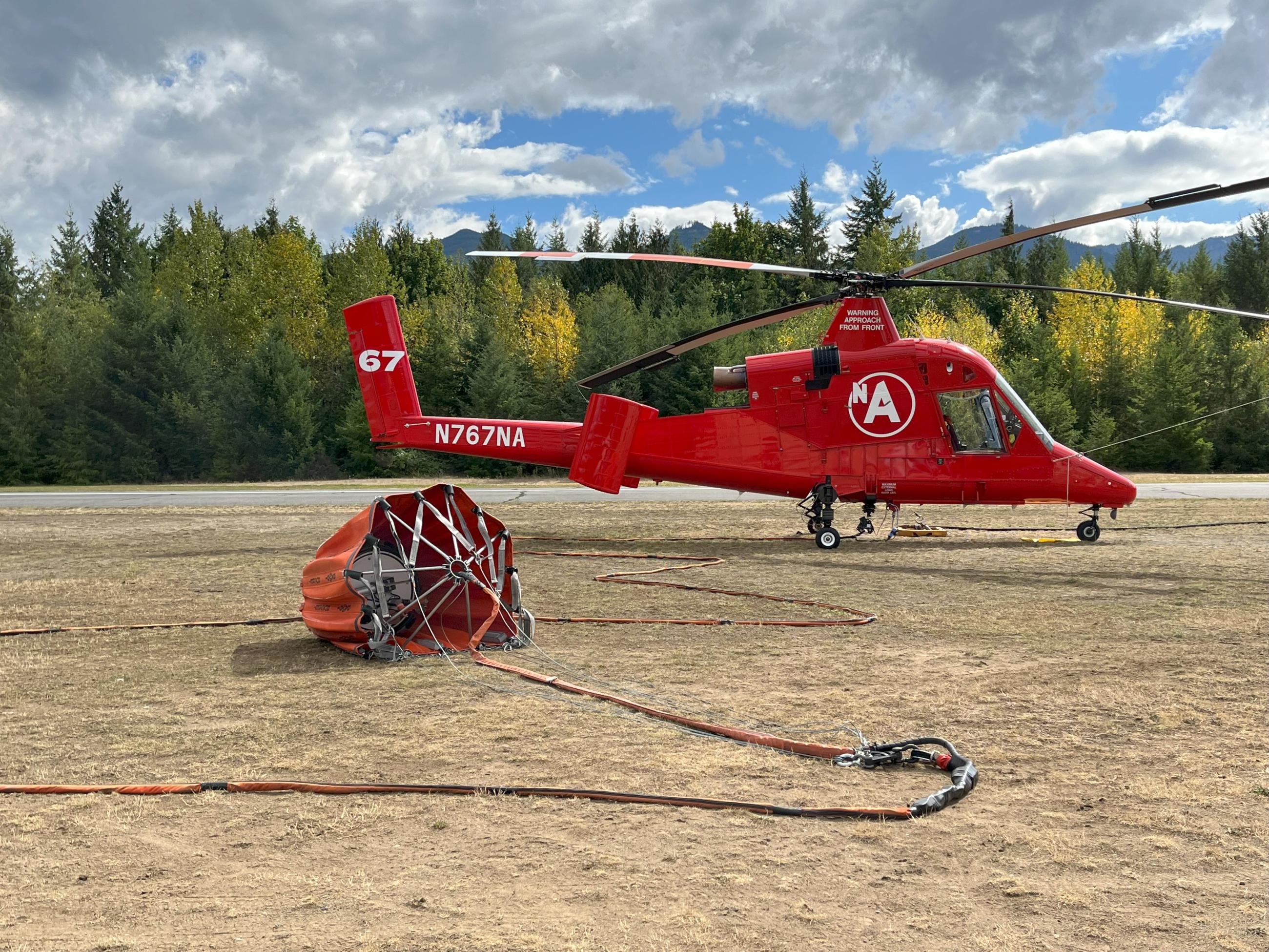 A helicopter sits on the ground with a large bucket for water drops attached and laid out in front of it.