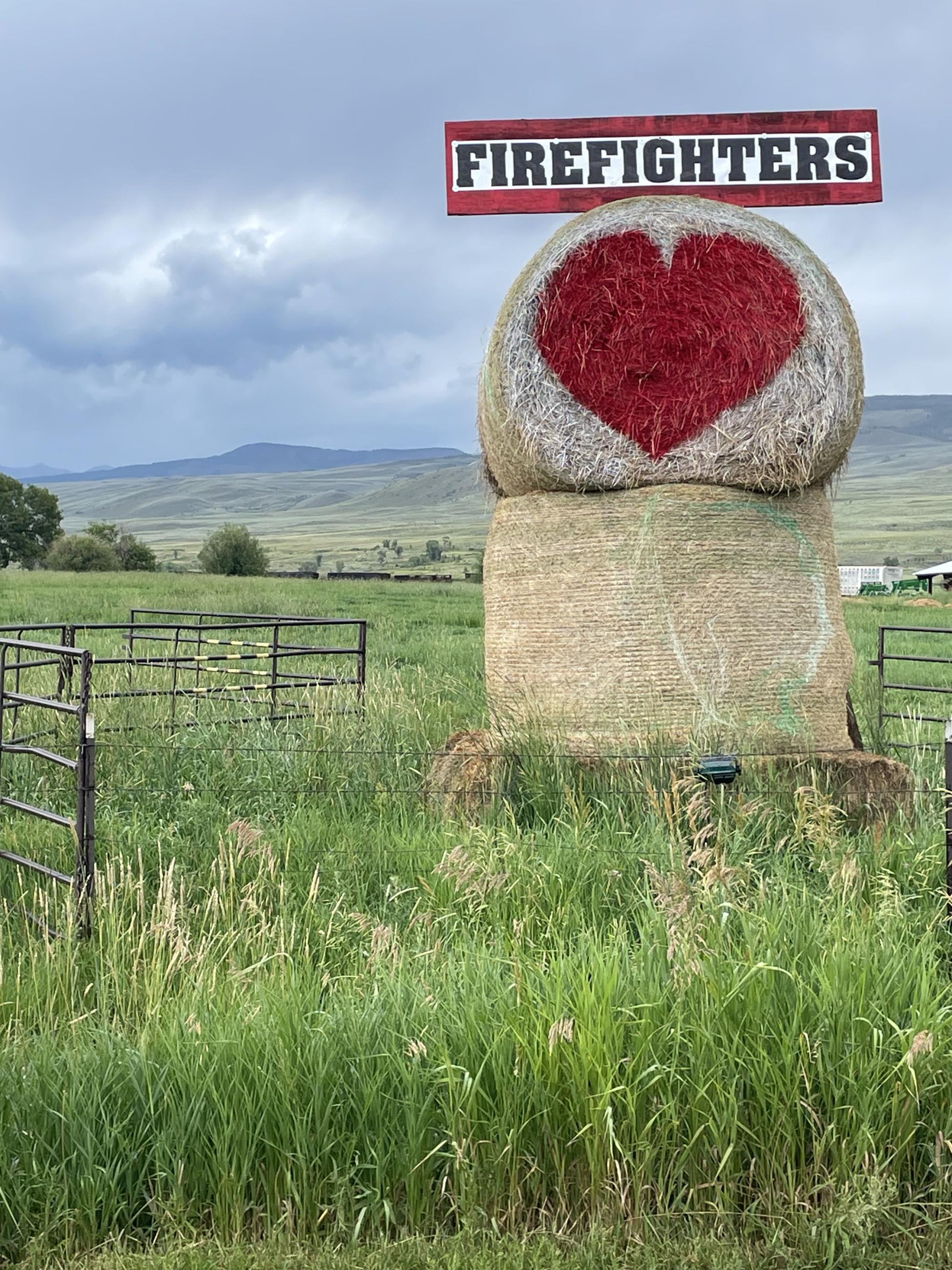 A round hay bale sitting on top of another bale with a big red heart painted in the center. A rectangular banner sign is mounted on top that says firefighters in big black block letters with a red border.