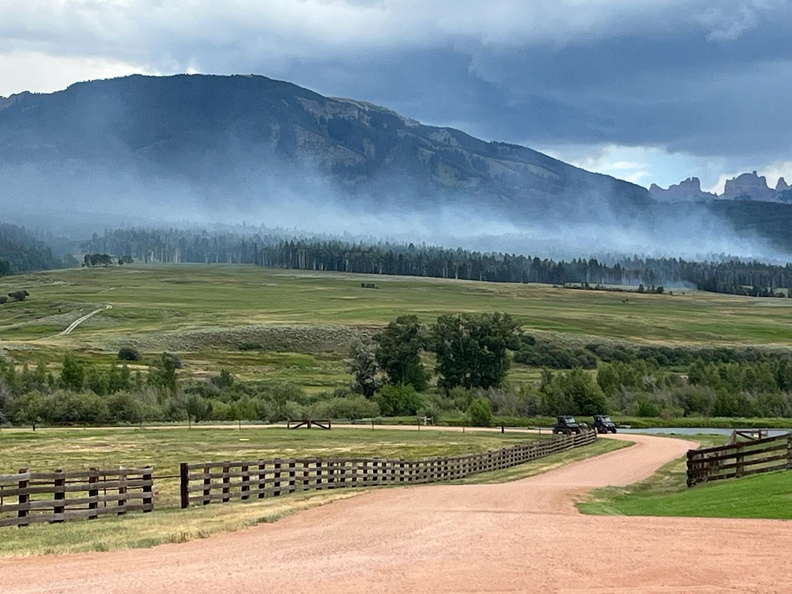 Light wispy smoke rising above the trees in front of a mountain as viewed from a ranch on the west side of the Lowline Fire.