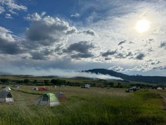 Scattered tents in the foreground at the Incident Command Post and smoke in the distance from the fire in Division A to the west. The sun is shining brightly through the clouds.