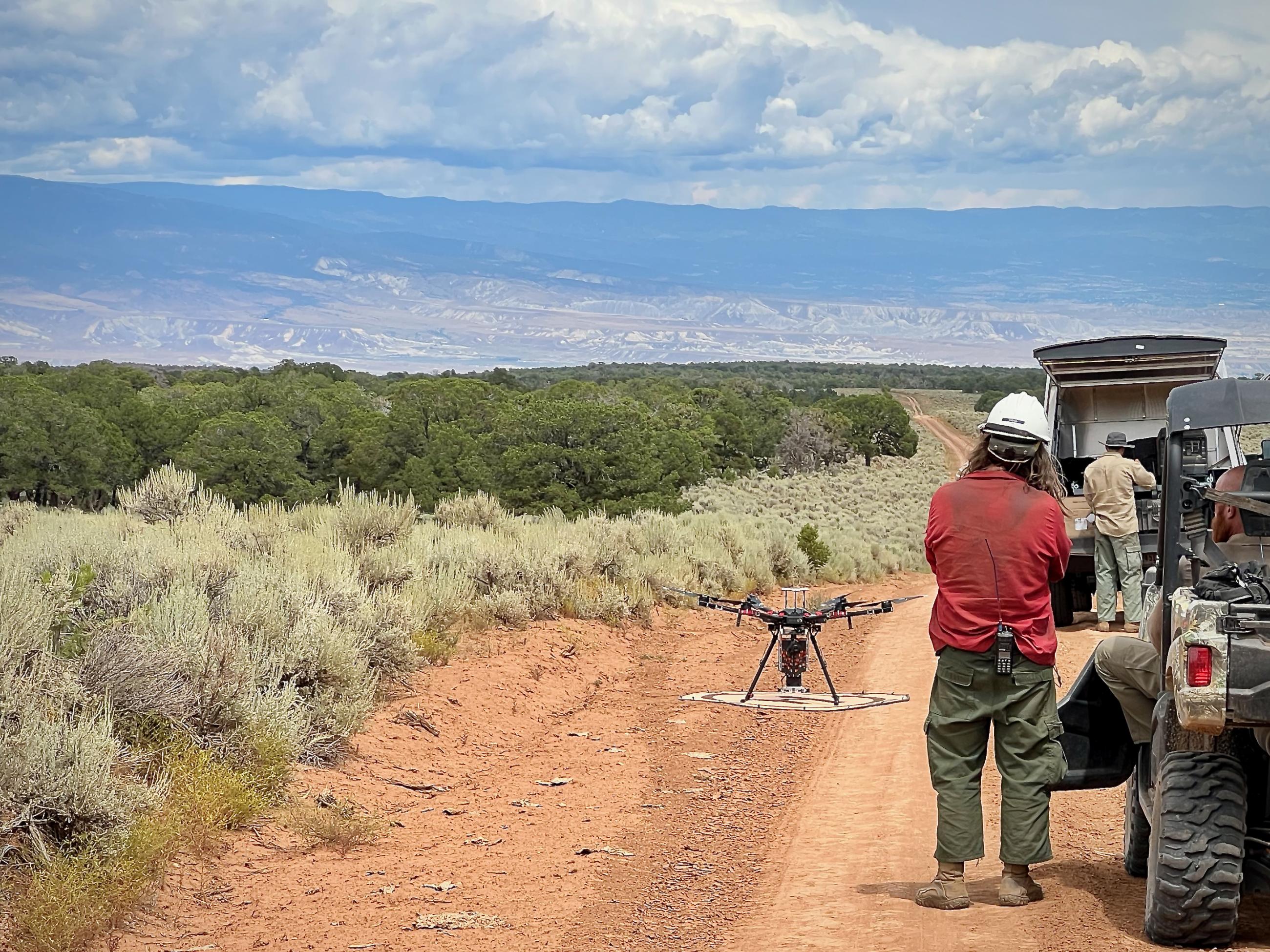 A firefighter wearing green pants, a red shirt, and a while helmet, with a radio clipped to his belt launches an unmanned aircraft system from a dirt road beside sage and juniper vegetation, with Grand Mesa in the background