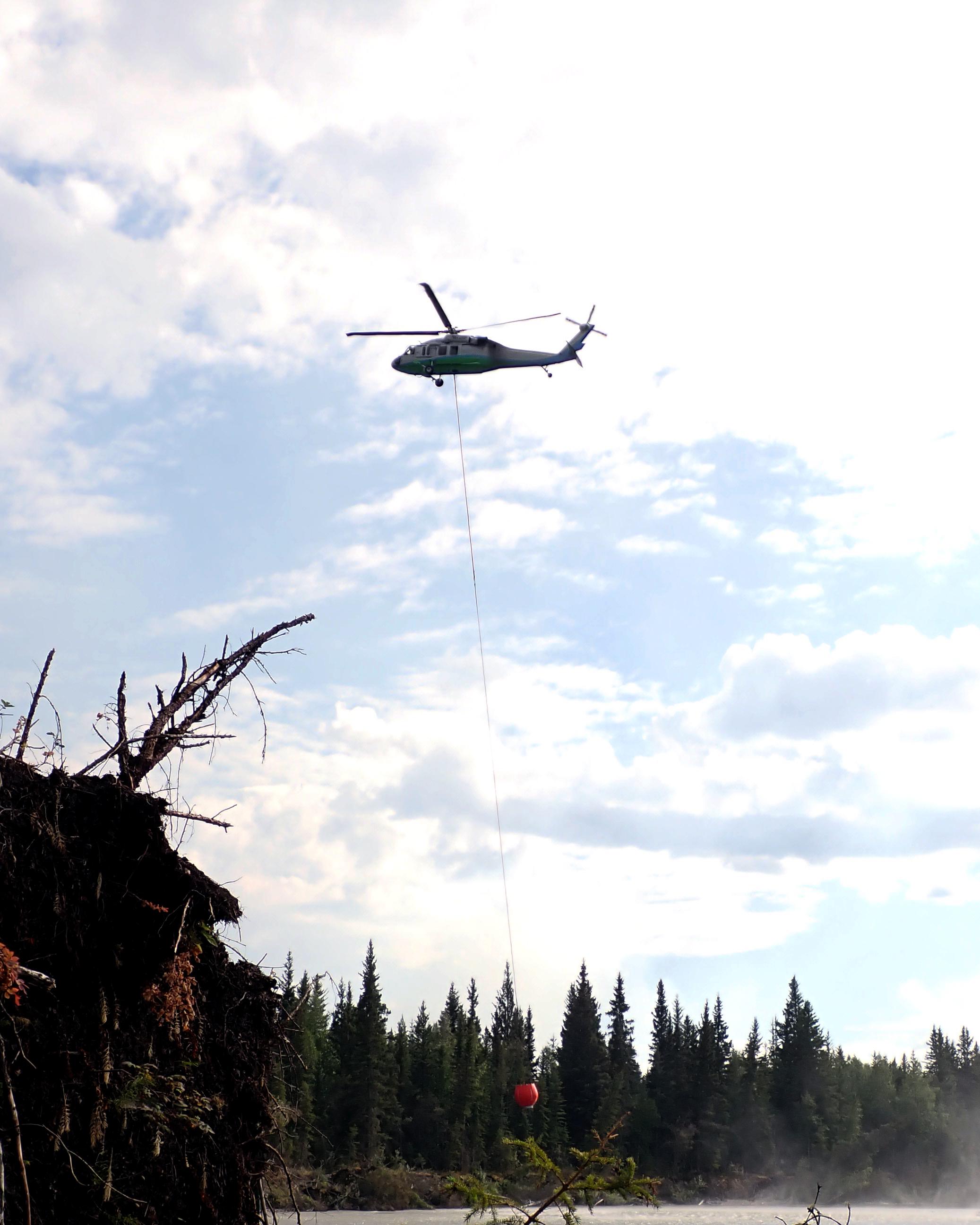 Photo of a Blackhawk helicopter silhouetted against a partly cloudy sky. The Blackhawk is using an orange bucket called a 'bambi bucket' to dip water out of the Teklanika River. The water will be dropped on hot spots on the Shores Landing Fire.