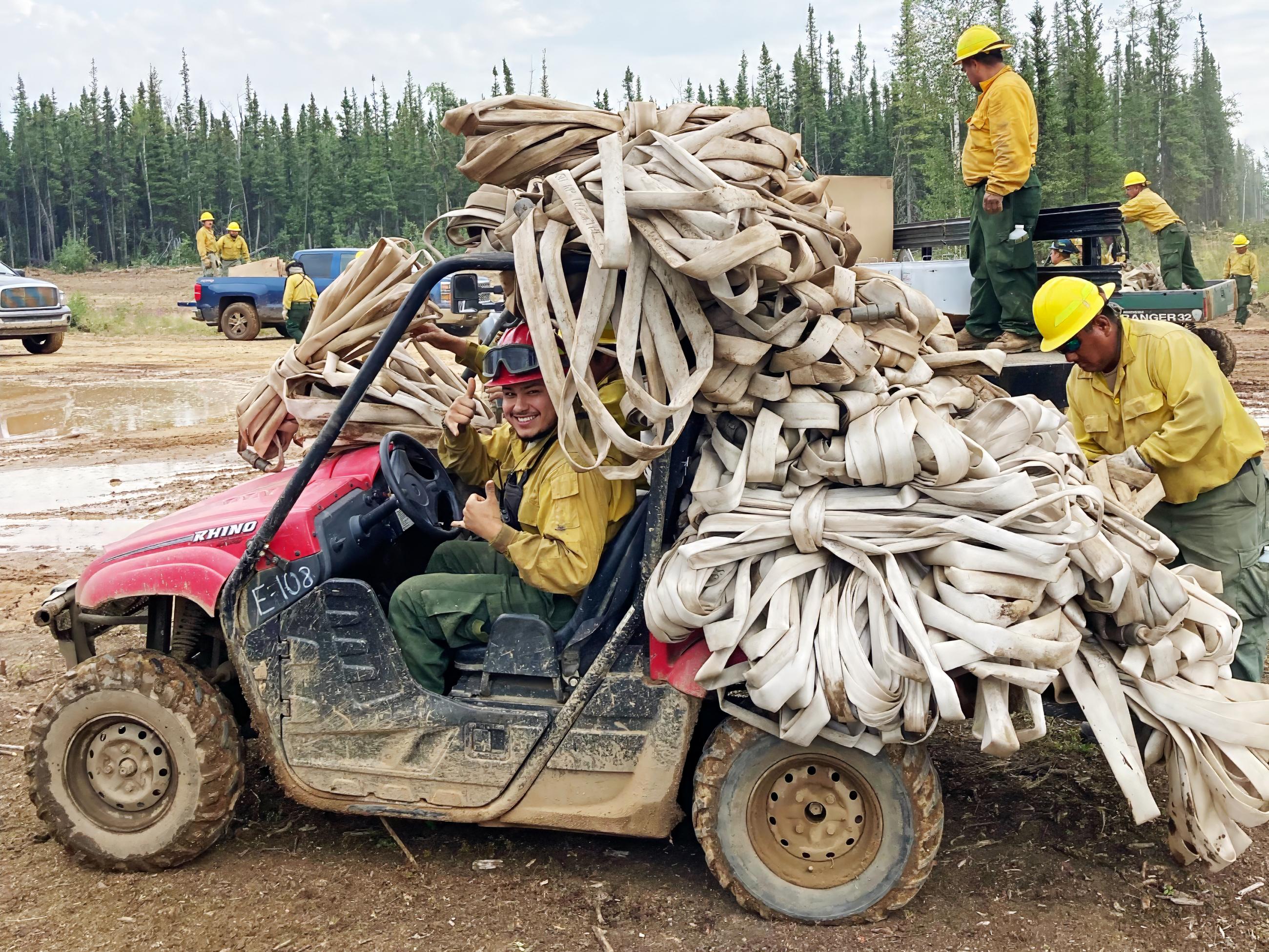 A UTV is loaded with hoses fills the entire frame the firefighter driving is smiling at the camera in yellow shirt and green pants as a second firefighter adds more hoses to the load in the back.