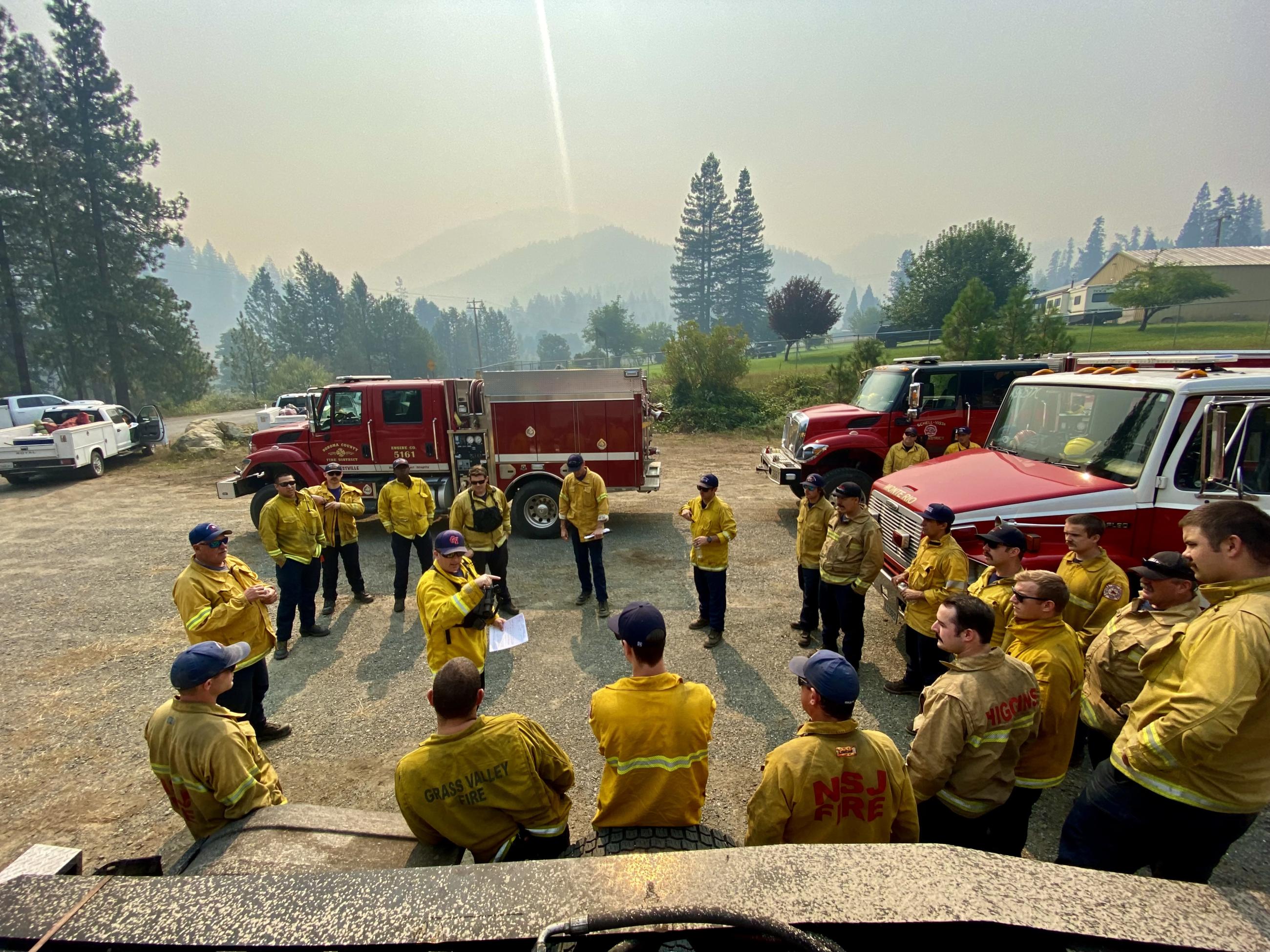 Image of firefighters standing in a circle with their engines behind for a briefing. Photo USDA Forest Service courtesy Daniel Ramey
