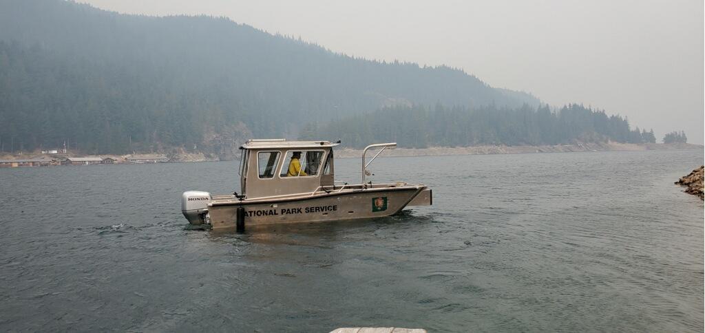 A photo of a boat with the words "National Park Service" on the side is idling in a large body of water with a smoke filled background.