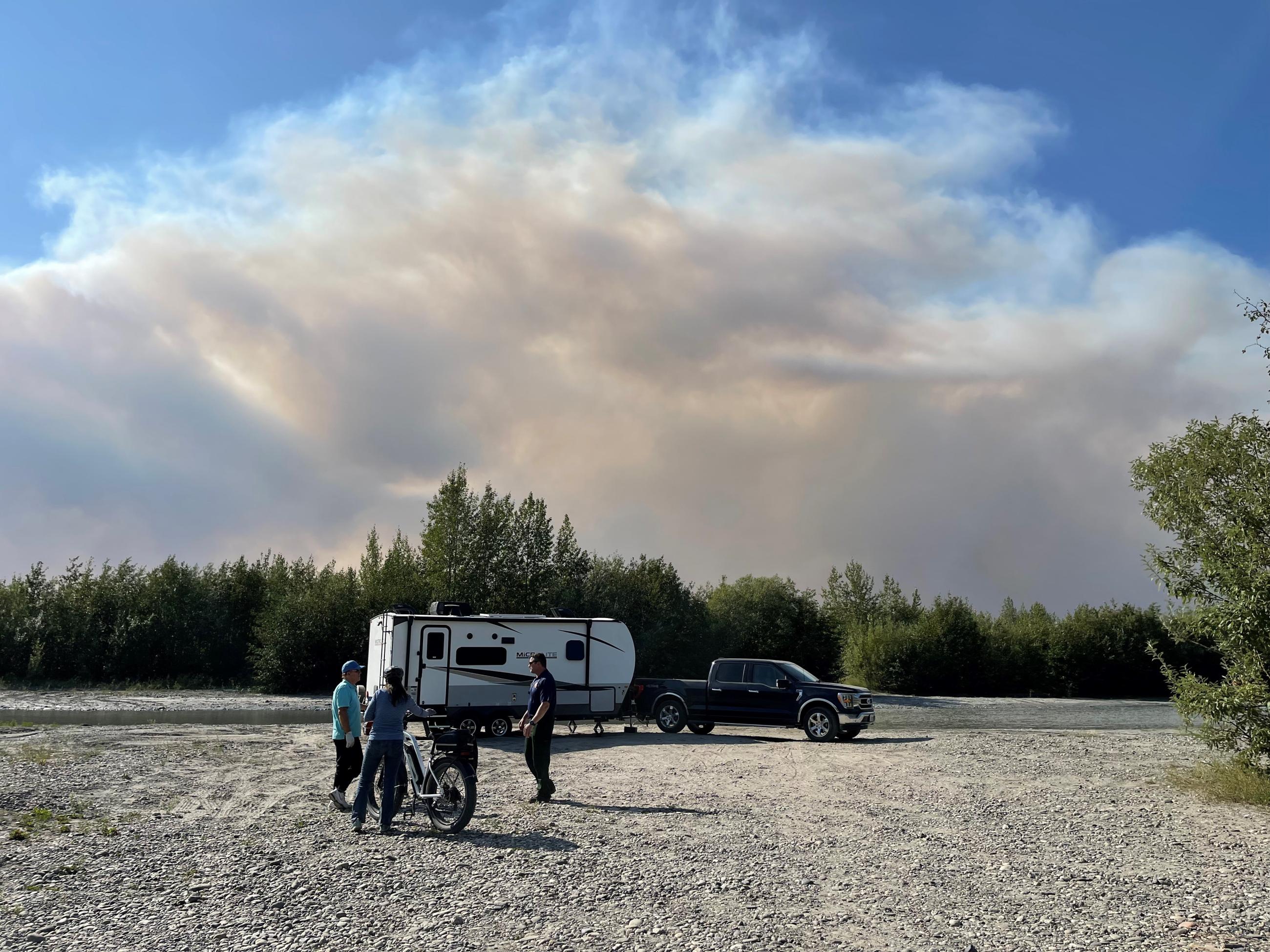 A large smoke column builds behind a camp trailer and pickup truck parked along the banks of a river while fire managers talk with campers in the foreground.