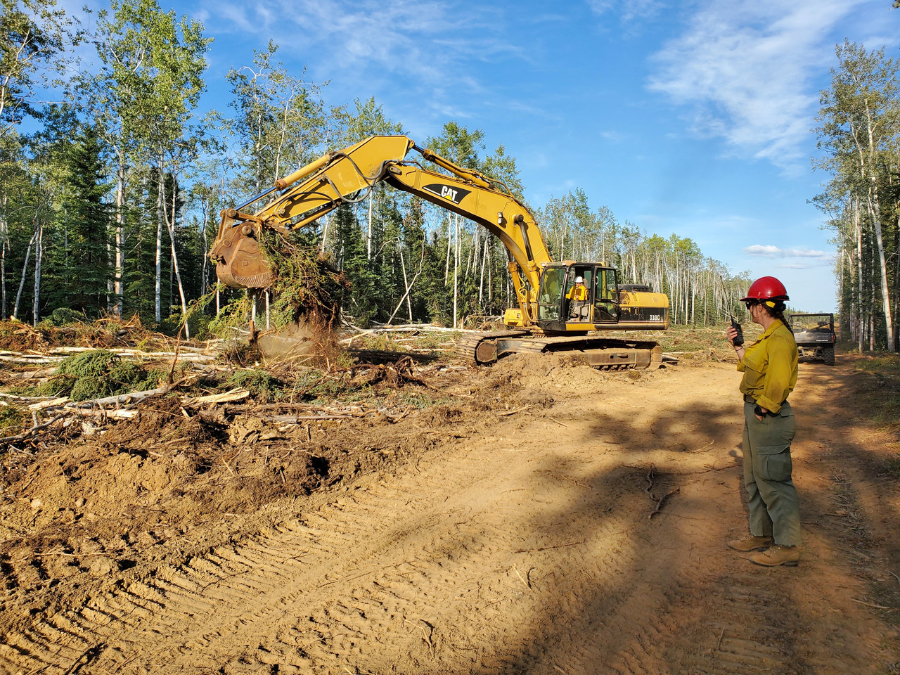 Photo showing an excavator moving vegetative debris and placing it on a firebreak. A resource advisor is talking to the heavy equipment operator by handheld radio.