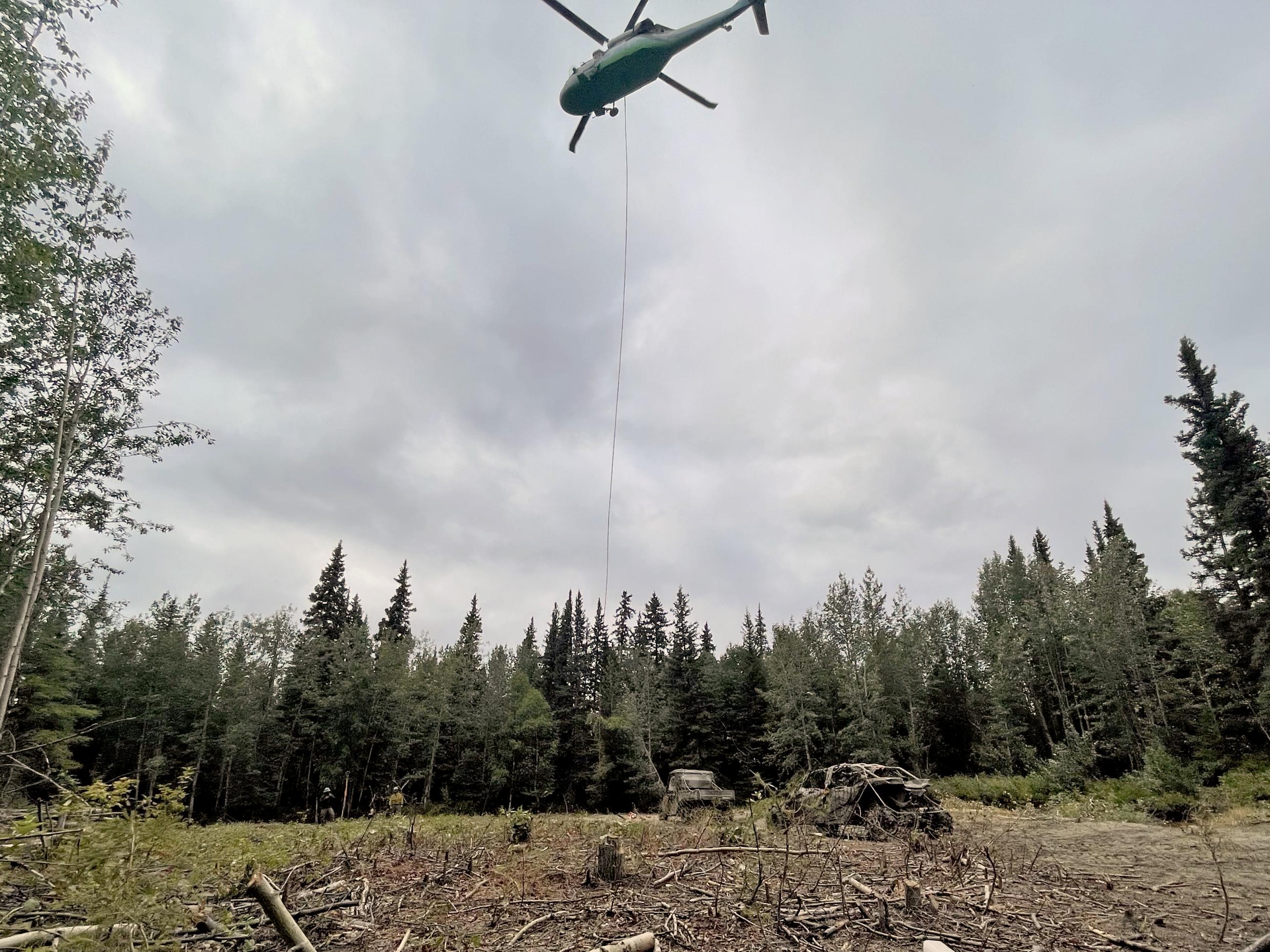 Helicopter hovers center of the photo with a long cable connected to a UTV in a webbing of large rope.