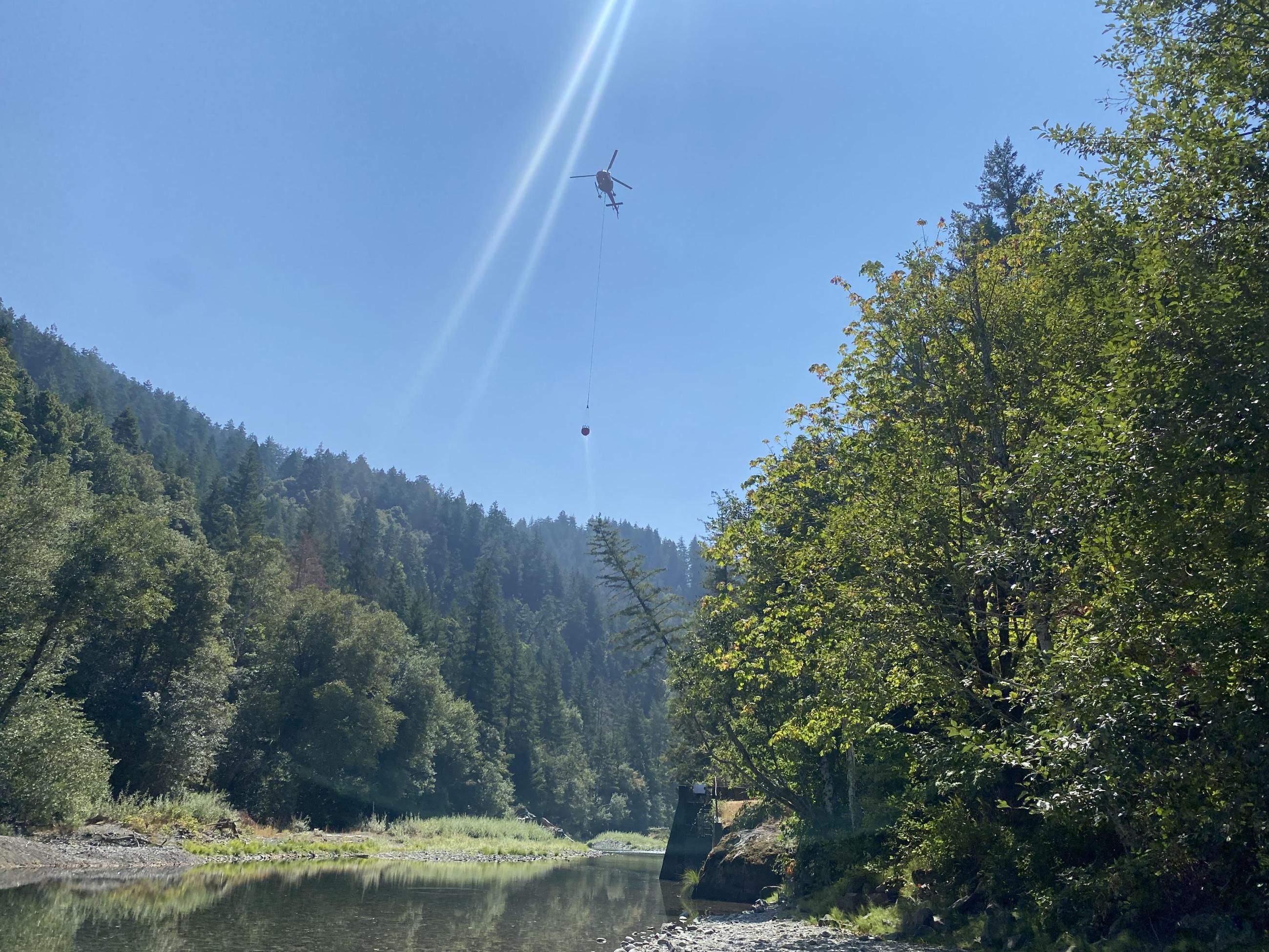 A helicopter soars through the sky with a bucket attached by a long a cable over the Elk River.