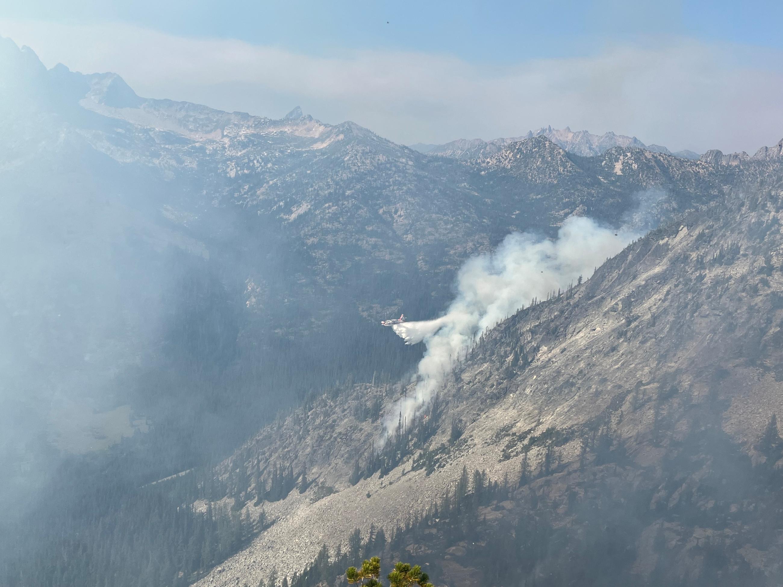 A scooper plane dropping water over a burning fire in a forested mountain range.