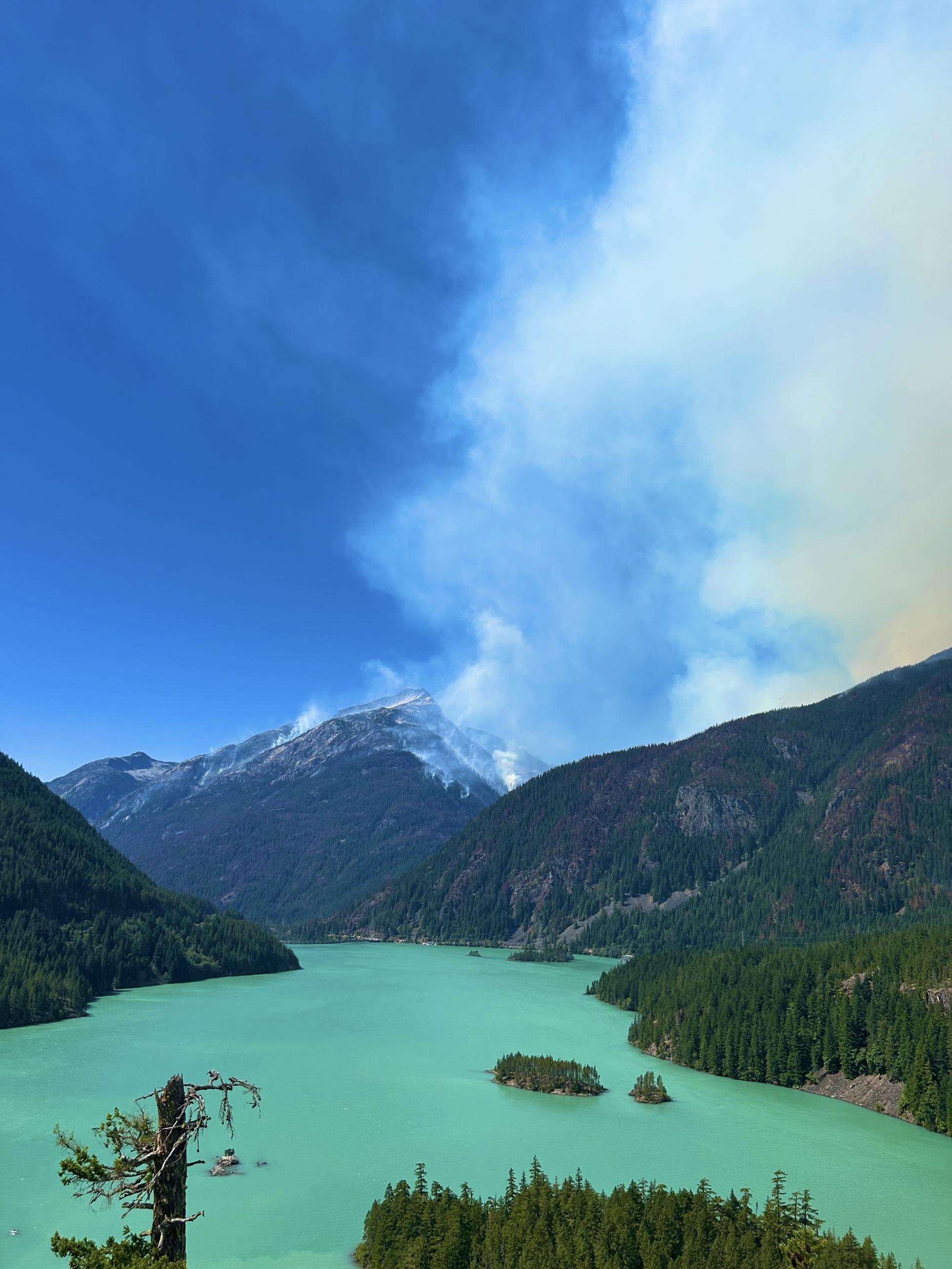 A photo of a lake in the foreground, with smoke rising from the hillside across the water.