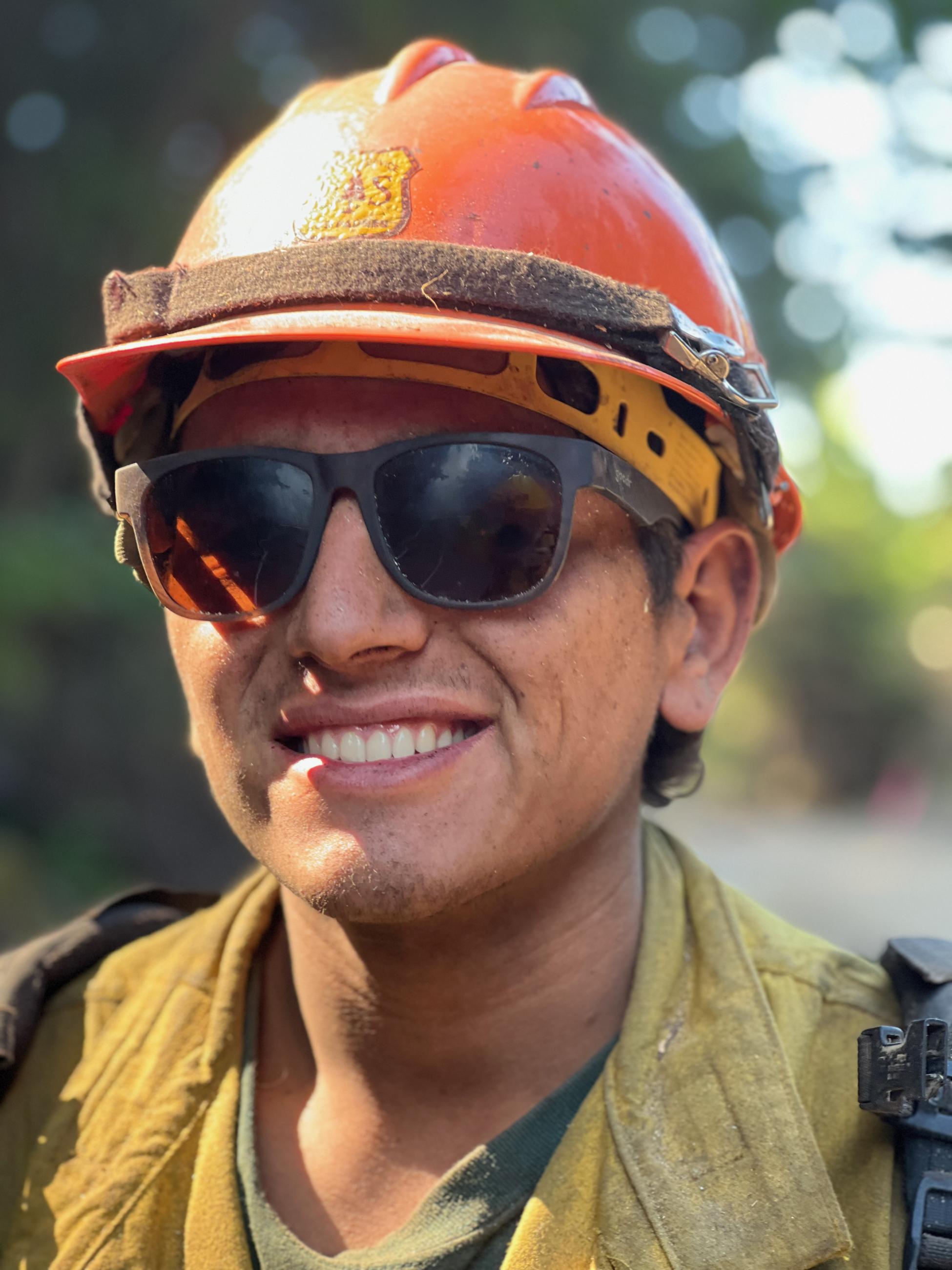 A portrait of a firefighter in a yellow shirt, orange helmet and wearing sunglasses.