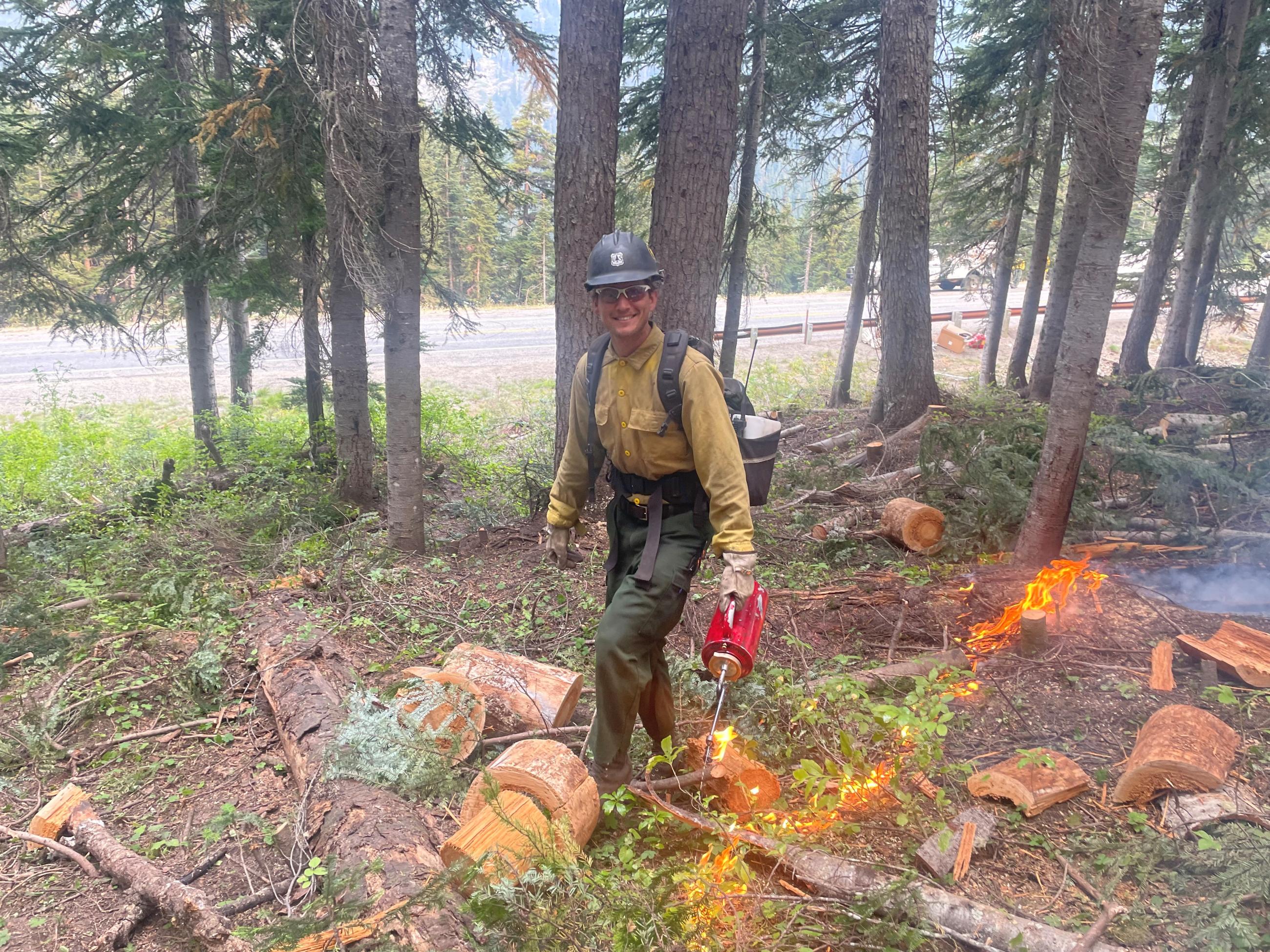 A wildland firefighter holding a red drip torch with active flame on the forest ground cover in the forest near highway 20