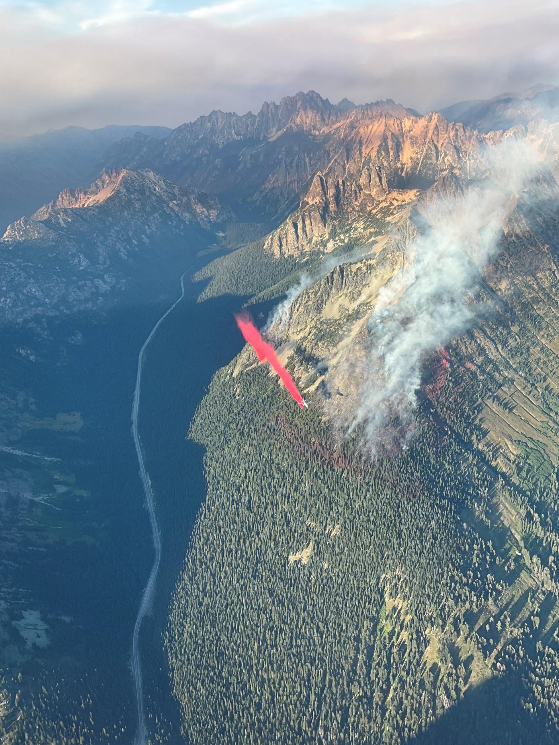 A large airtanker drops red fire retardant over a forested mountain range with fire smoke near scenic Highway 20.