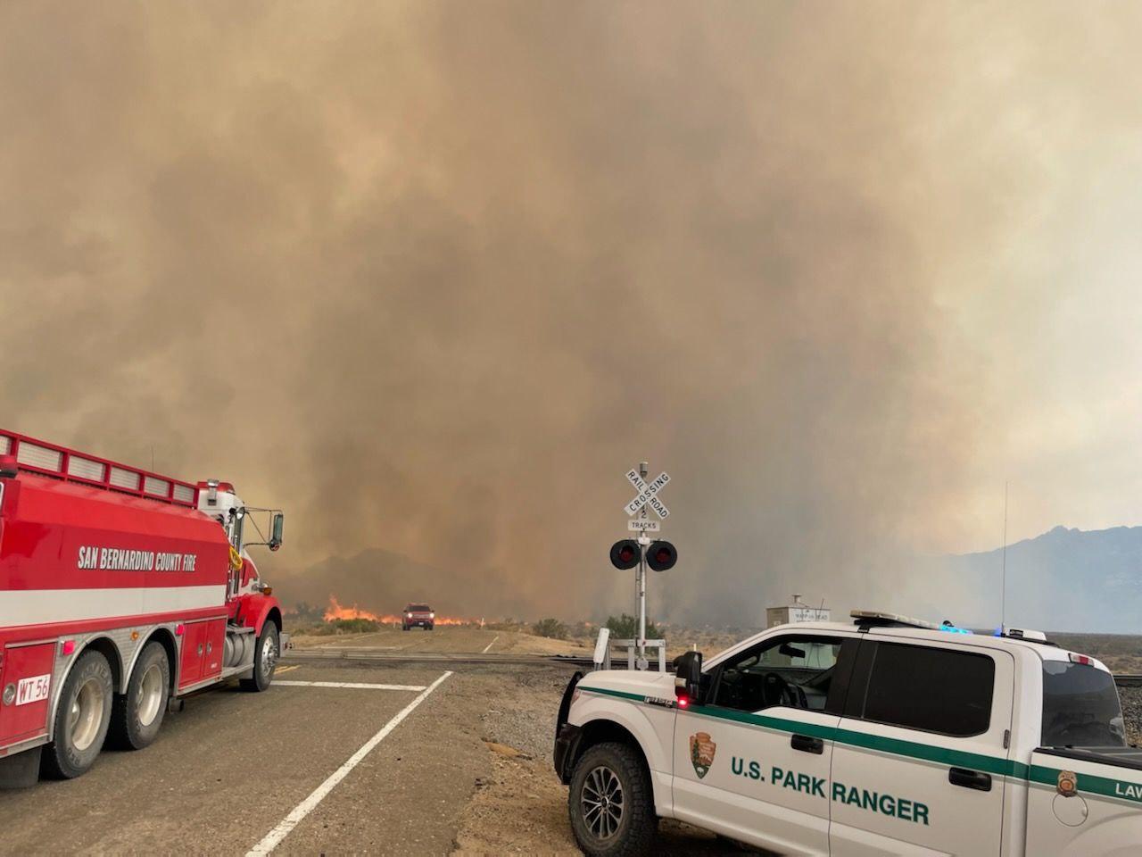 a red fire engine, and a National Park Service truck are parked near a railroad crossing sign.  Smoke and flames visable in the background.