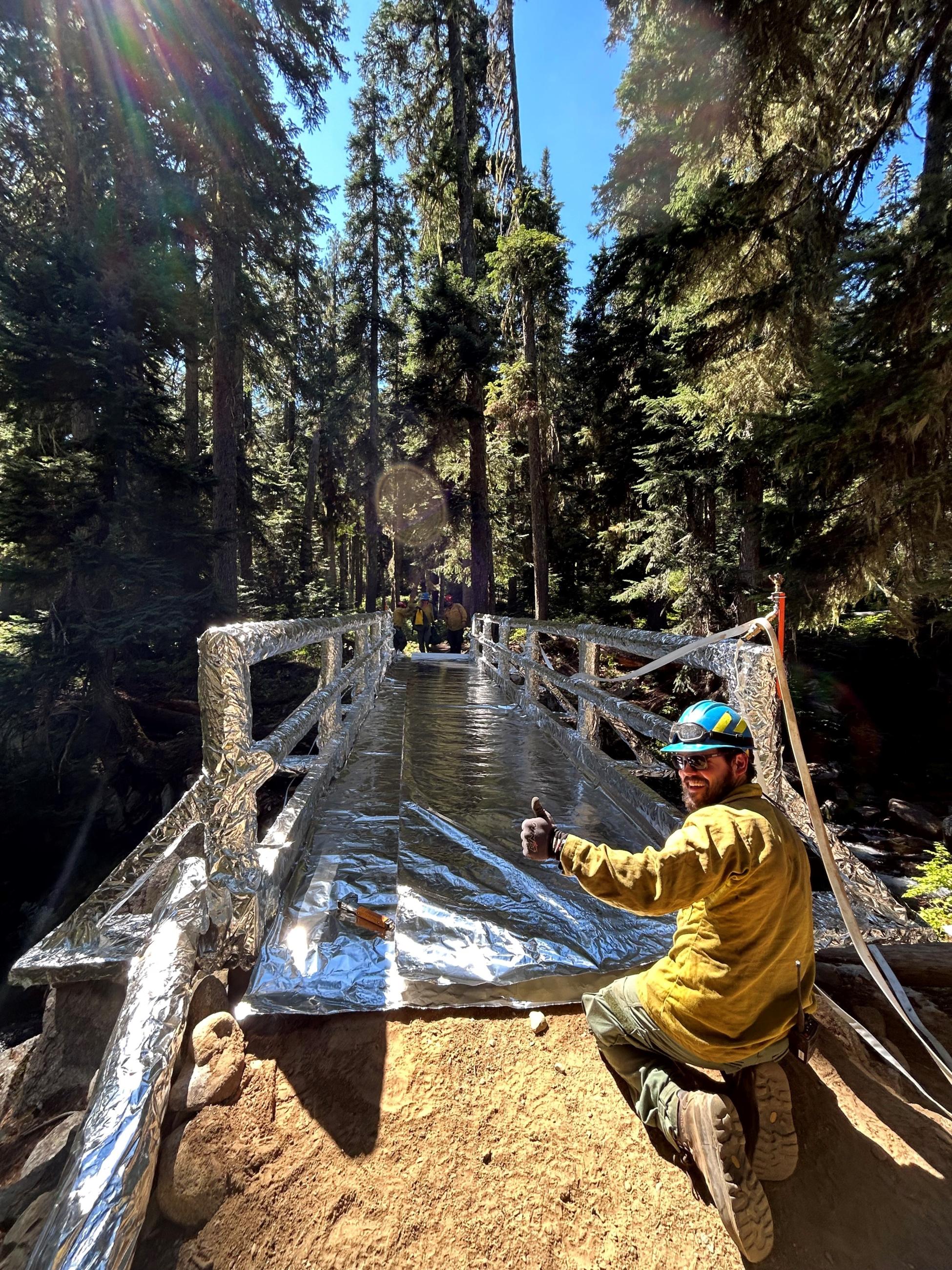 A forested area with Forest Service crews working to wrap a stock bridge in silver aluminized fire resistant structure wrap.